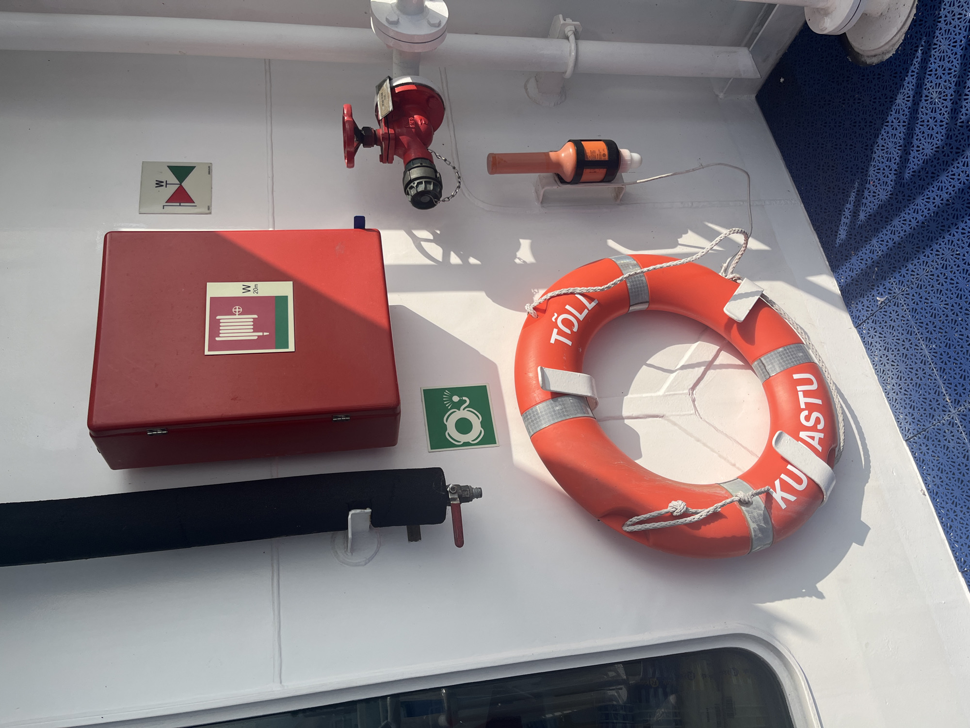 The image depicts a section of a white boat or ship's interior, likely in the city of Virtsu, Estonia, as suggested by the presence of nautical equipment. The scene is dominated by safety equipment mounted on the white wall. In the center, a bright orange life preserver with white lettering "TÖLL KUU ASTU" is hung on a white bracket. To the left of the life preserver, a green square sign with a white pictogram of a life preserver and water spray is visible. Above this, a thick black pipe is secured to the wall by a white bracket.
To the right of the pipe and above the green sign, a red fire hose cabinet with a metal latch is mounted on the wall. A square pictogram of a coiled hose with a spray nozzle and the text "W 20m" is affixed to the cabinet. Slightly to the right and above the fire hose cabinet, a square sign with a red and green triangular pictogram and the letter "W" is mounted on the wall.
Further to the right, a bright red fire hydrant with a wheel-shaped handle is connected to a white pipe. A small metal tag is attached to the hydrant. Below the hydrant, an orange signaling device, possibly a flare or distress signal, is mounted in a white holder. A thin wire extends from the device.
The white wall contrasts with the blue, patterned flooring visible in the lower portion of the image. Sunlight casts distinct shadows of the life preserver and other equipment onto the wall, indicating the presence of natural light. The overall environment suggests a functional, safety-focused area on a vessel.