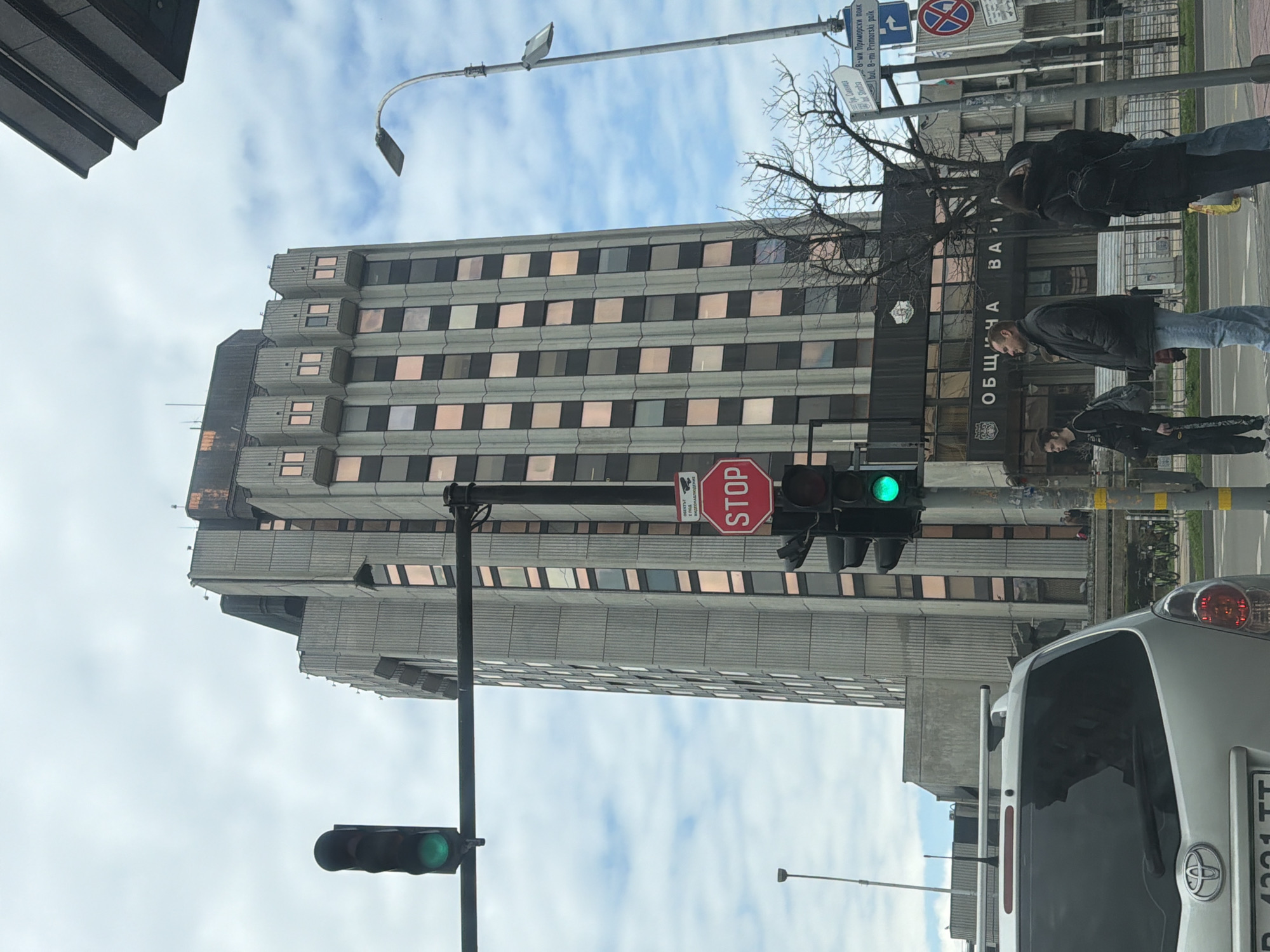 The image captures a street scene in Varna, Bulgaria. A tall, imposing building with a modern architectural style dominates the background, labeled with Cyrillic text "ОБЩИЧА" and "ВА" on its lower portion. Street signs indicate "8-ми Приморски полк" which translates to "8th Primorski Polk" in English. A stop sign and traffic lights are visible, with one traffic light displaying a green signal. Several pedestrians are walking on the sidewalk near the building. A portion of a silver Toyota with license plate "PB 4221 TT" is visible on the right side of the image, suggesting the photo was taken from inside a vehicle. The sky is overcast with scattered clouds.