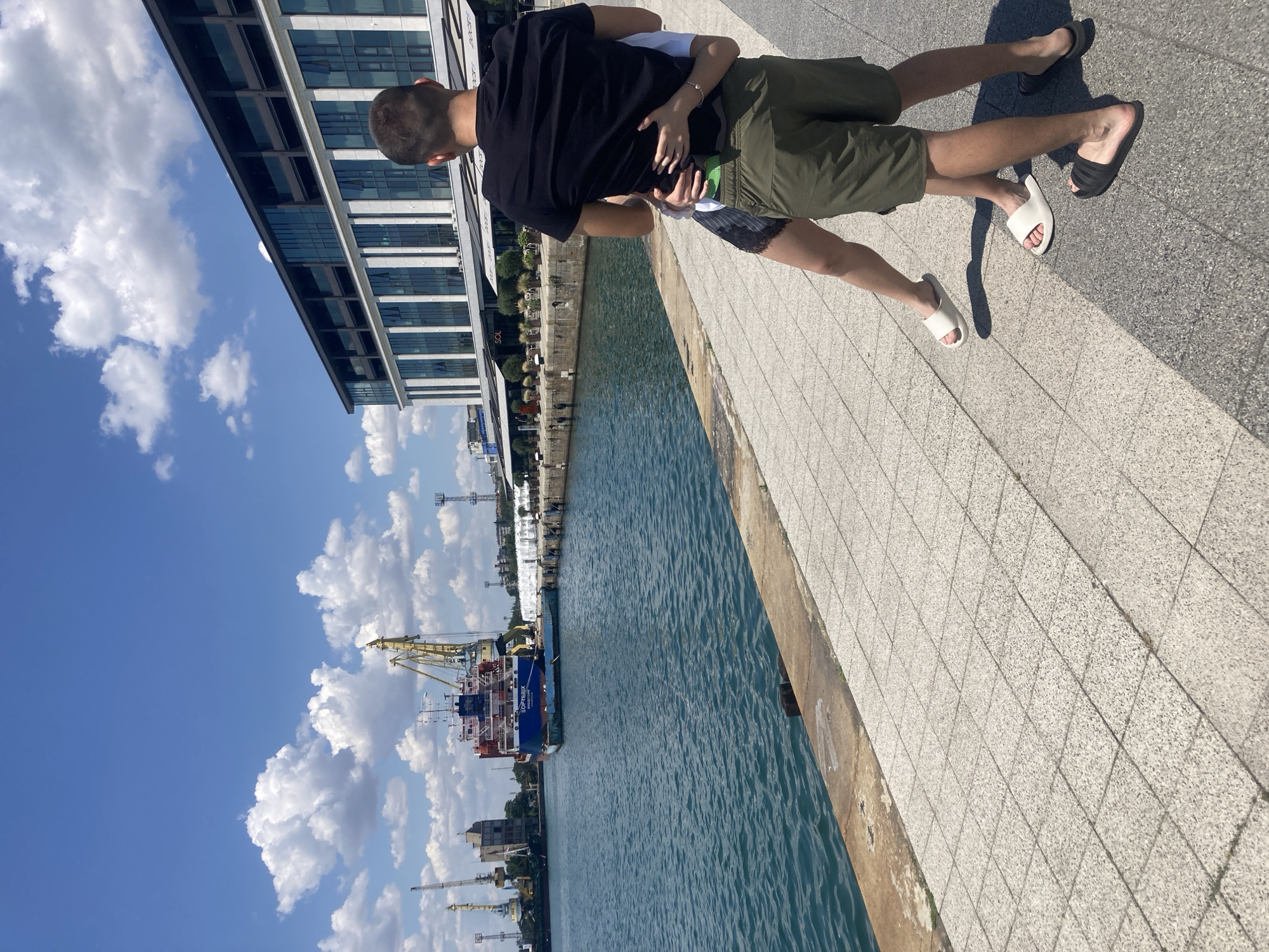 A couple embraces on a paved waterfront promenade in Burgas, Bulgaria. The man, wearing a black t-shirt and green shorts, holds the woman from behind. She wears a white t-shirt and dark shorts. The sea is to their left, with a large ship and industrial cranes visible in the background. To their right is a modern building with multiple floors and balconies, overlooking the promenade. The sky is blue with scattered white clouds. The promenade is paved with light-colored square tiles and bordered by a concrete edge.