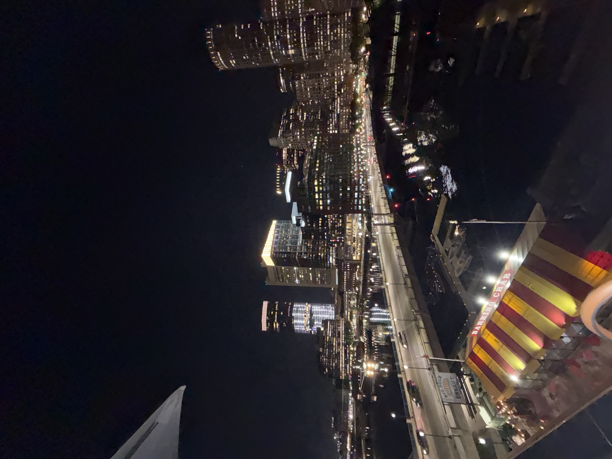 The image captures an aerial view of Boston at night, featuring lit-up skyscrapers along a waterway. A bridge stretches across the water, with vehicles moving along it. In the foreground is the "Barking Crab" restaurant, identifiable by its red and yellow striped awning and the sign that reads "Big Dice Energy." A small portion of a white, sail-like structure is visible in the lower left corner.