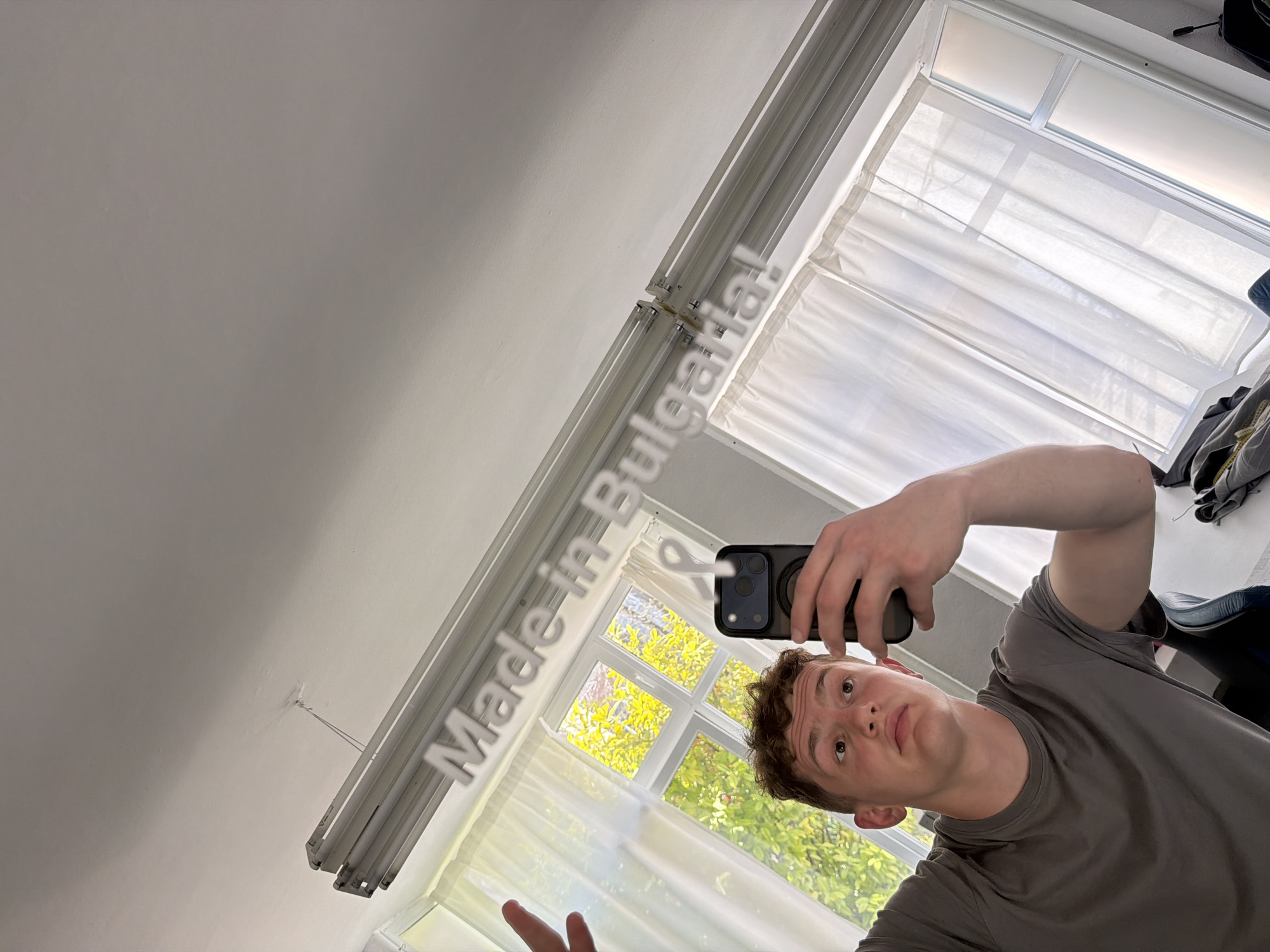 A fair-skinned young man takes a selfie in a room with a window. He holds a black smartphone in his right hand, which partially covers his face. He has brown curly hair and wears a grey t-shirt. The room has a window with white curtains and a view of green trees and foliage outside, suggesting it is daytime. Fluorescent lights are affixed to the ceiling. In the background, there is a desk with a chair, and some clothes scattered on the surface. The phrase "Made in Bulgaria!" is digitally overlaid in white, block letters in the image. The precise location within the city of Varna, Bulgaria, is impossible to determine solely from visual elements.