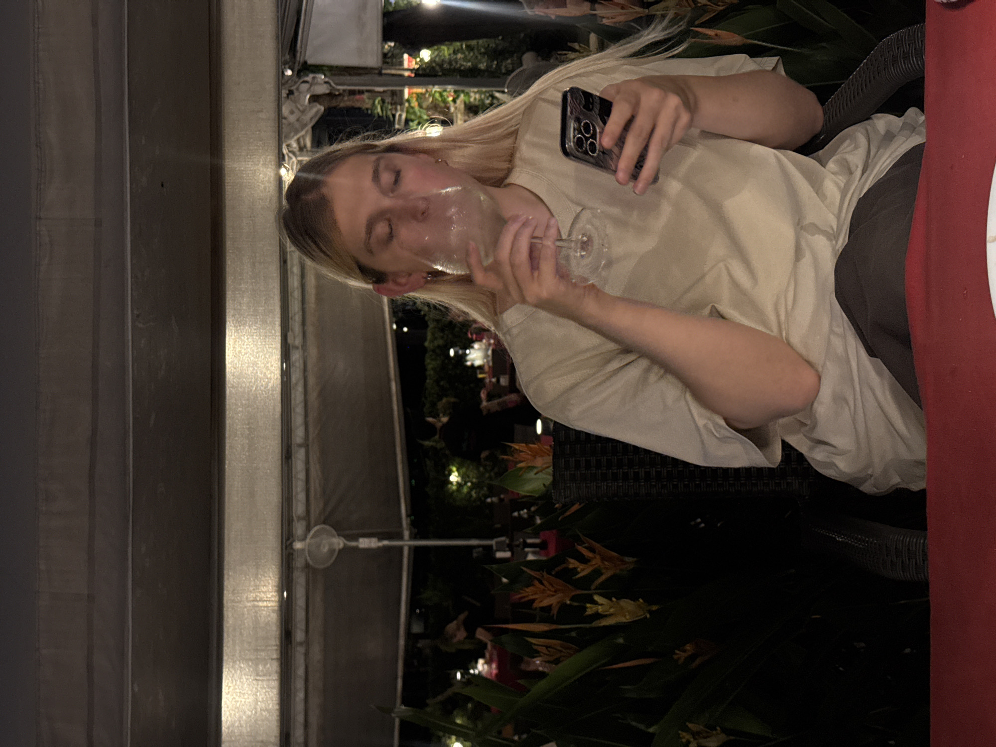 The image shows a young person with blonde hair at an outdoor dining establishment. The person is wearing a light-colored t-shirt and dark pants. They are holding a wine glass in their right hand and a smartphone in their left hand. The person is drinking from the wine glass, and their eyes are closed. The background is dark, with some artificial lights visible, suggesting it is nighttime. There are tropical plants and foliage in the background, which, along with the dim lighting and outdoor setting, hint at a warm climate. The person appears relaxed or perhaps in a moment of enjoyment. The image is oriented vertically, with the person positioned on the left side of the frame. A red tablecloth or surface is partially visible on the right side of the image. The overall impression is a casual evening out.