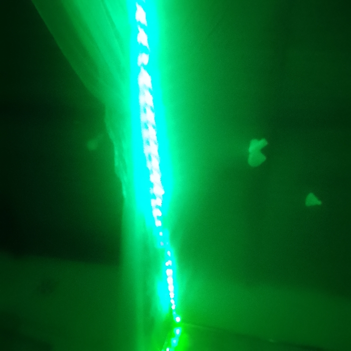 A vertical chain of bright, cyan-colored lights is set against a dark green background. The string of lights appears slightly blurred. Scattered in the green space around the light string are faint, distorted, ghostly shapes. There is a dark, undefined form on the left side of the image. The overall effect creates an eerie or otherworldly atmosphere.