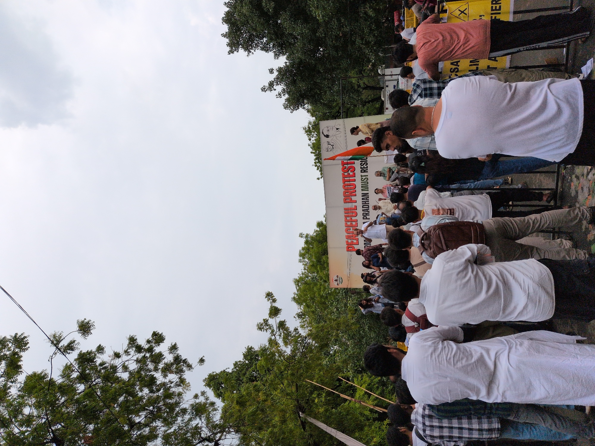 A crowd of people are gathered for a peaceful protest in New Delhi, India. The image captures a side view of the event, with a banner prominently displayed that reads "PEACEFUL PROTEST PRADHAN MUST RESIGN". An Indian flag is also visible. The sky is overcast, suggesting a cloudy day. The people appear to be predominantly male, dressed in casual attire. Some individuals are looking towards the banner, while others are engaged in conversation. The overall mood seems to be one of focused participation in the demonstration. Trees and foliage frame the scene, indicating an outdoor setting, possibly a street or public square. The presence of barriers suggests a controlled or organized gathering.