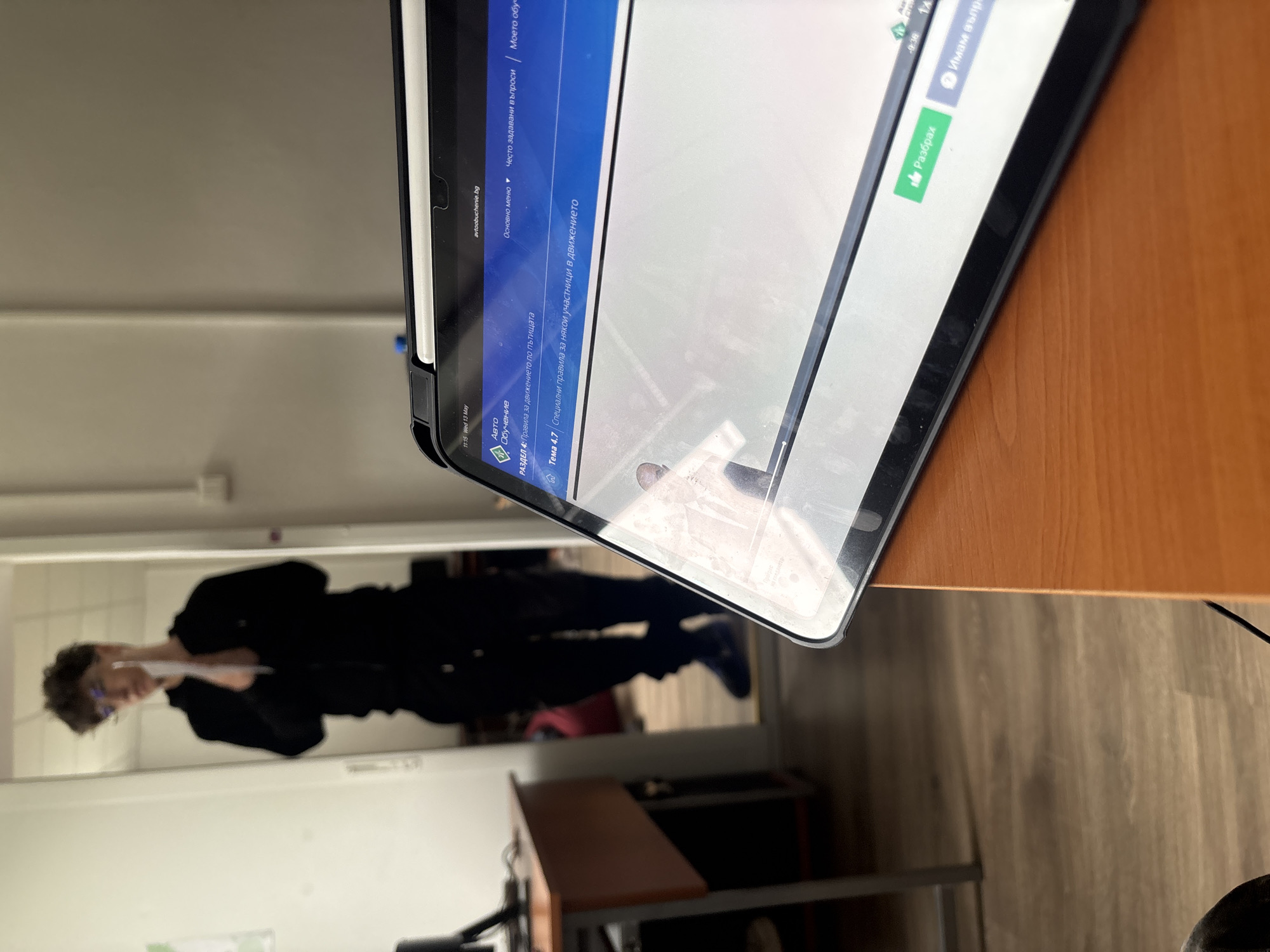 The image shows a tablet displaying a website related to driving education ("avtoobuchenie.bg"). The screen shows text in Bulgarian. In the reflection on the tablet's screen, a person in dark clothing and glasses can be seen standing in a room, possibly holding a document. The tablet is resting on a wooden desk with a cable running along the right side. The room appears to be indoors with a desk, chair and mirror. Without additional information, the location in Troyan, Bulgaria cannot be confirmed.