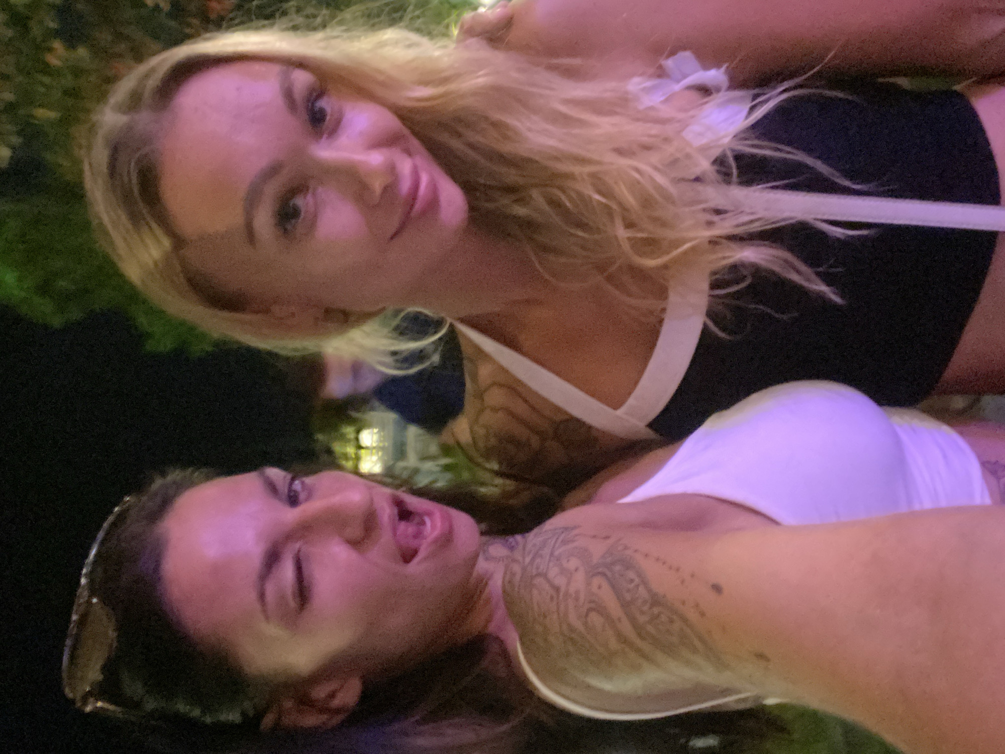 Two women are taking a selfie in an outdoor setting at night. The woman on the left is winking and has her tongue sticking out slightly. She is wearing a white top and has tattoos on her arm. The woman on the right has blonde hair and is smiling. She is wearing a black top with white trim. The background appears to be a street or an outdoor seating area with some greenery. The lighting suggests it is nighttime. The scene is likely in the city of Mũi Né, Vietnam, known for its beaches and vibrant nightlife.