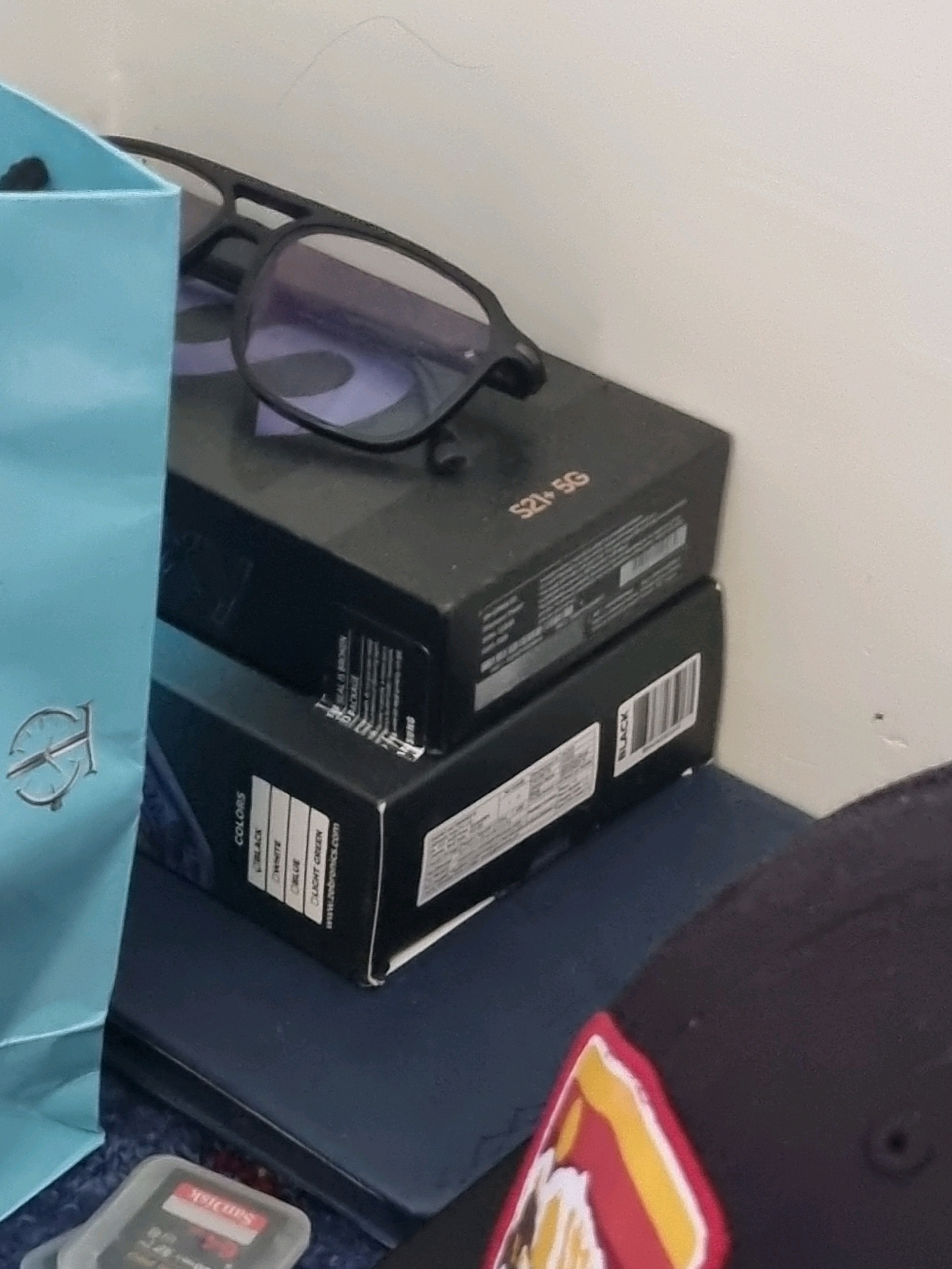 In Manigam, India, on a shelf, two black boxes are stacked, one atop the other. The top box displays "S21+ 5G" and has a sticker with text and a barcode. On the bottom box, a label reads "COLORS" with options like "BLACK," "WHITE," "BLUE," and "LIGHT GREEN," followed by a website address. To the left, a light blue shopping bag with a metallic emblem is visible. In the foreground, resting on a dark surface, is a clear plastic case containing a SanDisk memory card. Further to the right, a portion of a dark baseball cap with a red, yellow, and white emblem is present. Glasses with black frames are perched on top of the upper box.
