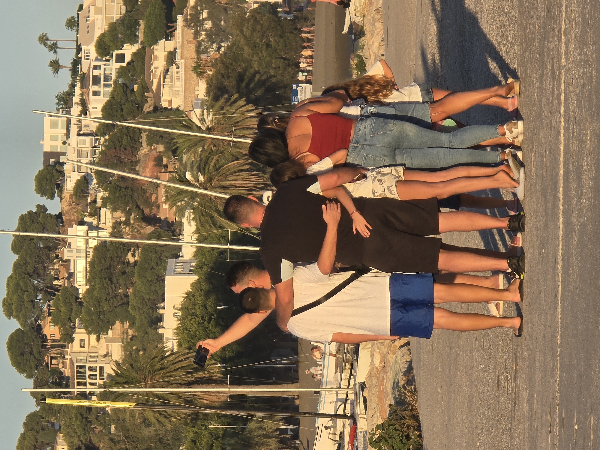 A group of six people, including at least two adults and four children, are gathered on a paved walkway near a harbor with sailboats. One adult male is holding a smartphone up to take a selfie of the group. He has his left arm around another male, who is wearing a white t-shirt and blue shorts. Another adult male in a black t-shirt and black shorts has his arms around the two males in front of him. A woman in a red tank top and jeans is standing next to the man in black, with her arm around a younger girl. Another younger girl stands beside them. The background features a hillside covered with trees and white buildings, characteristic of a Mediterranean coastal city like Málaga, Spain. Sailboats are docked in the water to the left of the group, and a beach with people is visible in the distance on the right. The lighting suggests it is late afternoon or early evening.
