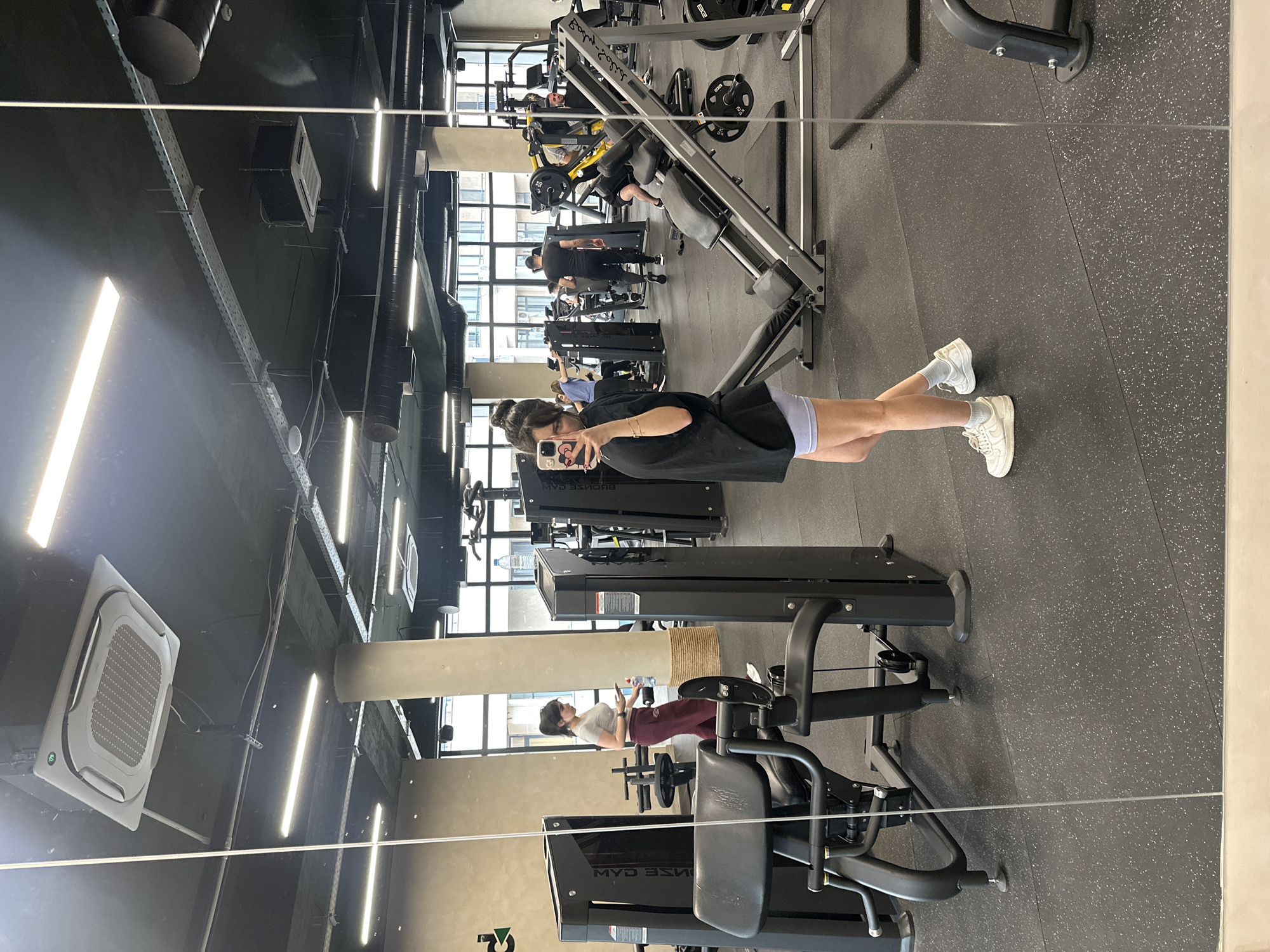 The image depicts a woman taking a selfie in a gym. The gym appears to be located in an urban area, possibly the city of Yerevan, Armenia, given the query parameters. The woman is dressed in athletic attire, including a black t-shirt, shorts, white socks, and sneakers. She is holding her phone in her right hand, presumably to capture her reflection in a large mirror. The gym is equipped with various weight machines and fitness equipment. Other individuals can be seen in the background, engaged in their workouts. The gym has a modern interior with exposed ceilings, fluorescent lighting, and air conditioning units.