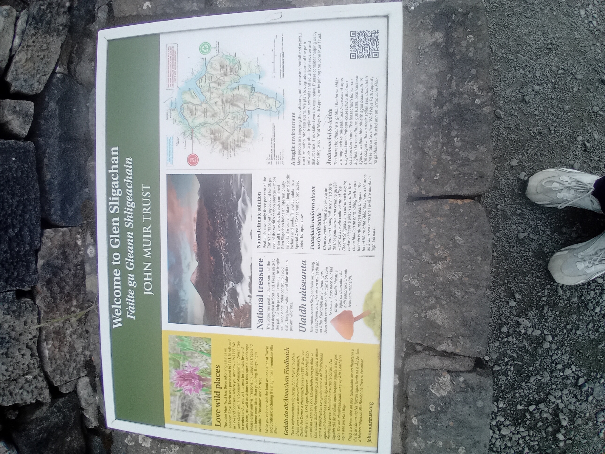 The image shows a sign at Glen Sligachan on the Isle of Skye, United Kingdom. The sign, titled "Welcome to Glen Sligachan" and "Fàilte gu Gleann Shligeachain" with "JOHN MUIR TRUST" below, is a white framed informational panel with a green border. It features text and images divided into sections. On the left, under the heading "Love wild places", there is a photograph of a pink flower and text in English and Gaelic. Below this is the website address "johnmuirtrust.org". In the center, under "National treasure", is a photograph of a mountain landscape and text in English and Gaelic, accompanied by an image of a mushroom. To the right, the top section is a map of the area with highlighted routes. Below the map, under "A fragile environment", there is text in English and Gaelic, including a QR code and the John Muir Trust logo. The sign is placed outdoors, with the base resting on a stone and gravel surface. The feet of a person wearing athletic shoes are visible at the bottom of the image, suggesting the sign is at ground level. The background is a dry stone wall.