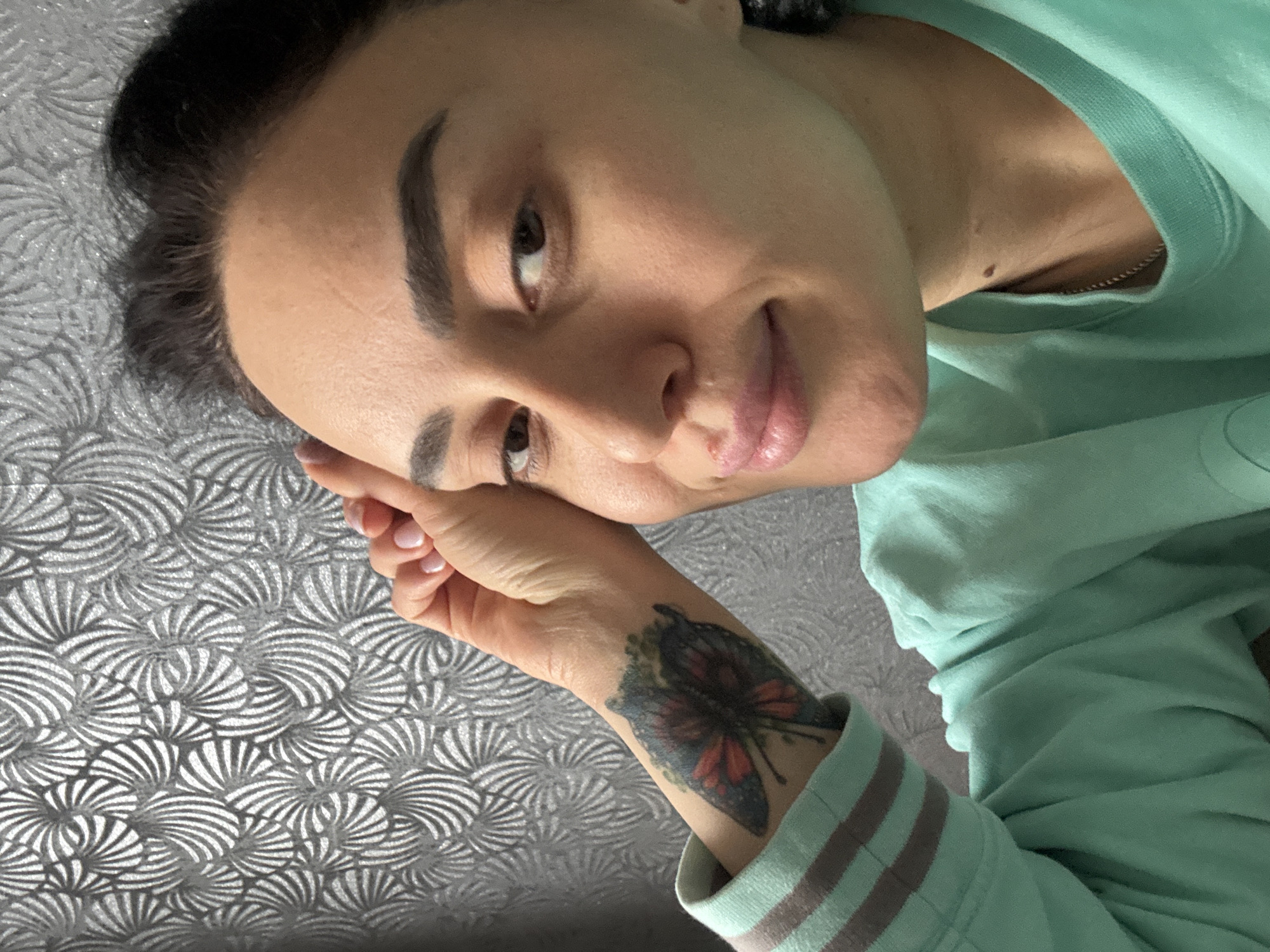A close-up, high-angle self-portrait features a woman in an indoor setting, likely in Tbilisi, Georgia. She looks directly at the camera with a calm, soft expression, her head slightly tilted and resting on her left hand. Her dark hair is pulled back, and she has well-defined, dark eyebrows. Her complexion is clear, showing faint lines on her forehead and around her eyes, and her lips are slightly parted in a subtle smile. A small, dark mole is visible on her neck, just below her jawline.
She is dressed in a light mint green long-sleeved top, possibly a sweatshirt, which features grey horizontal stripes on the cuffs. A thin silver chain necklace can be seen peeking from beneath the collar of her top. Her left hand is prominent, with light-colored, neatly manicured fingernails. On her left wrist, a distinct butterfly tattoo is visible, incorporating shades of red, black, and blue.
The background features a textured, light gray or silver wallpaper with a repeating, intricate fan or shell-like pattern. The lighting appears soft and natural, suggesting the image was taken during the day. There is no visible text in the image. The overall impression is relaxed and intimate.