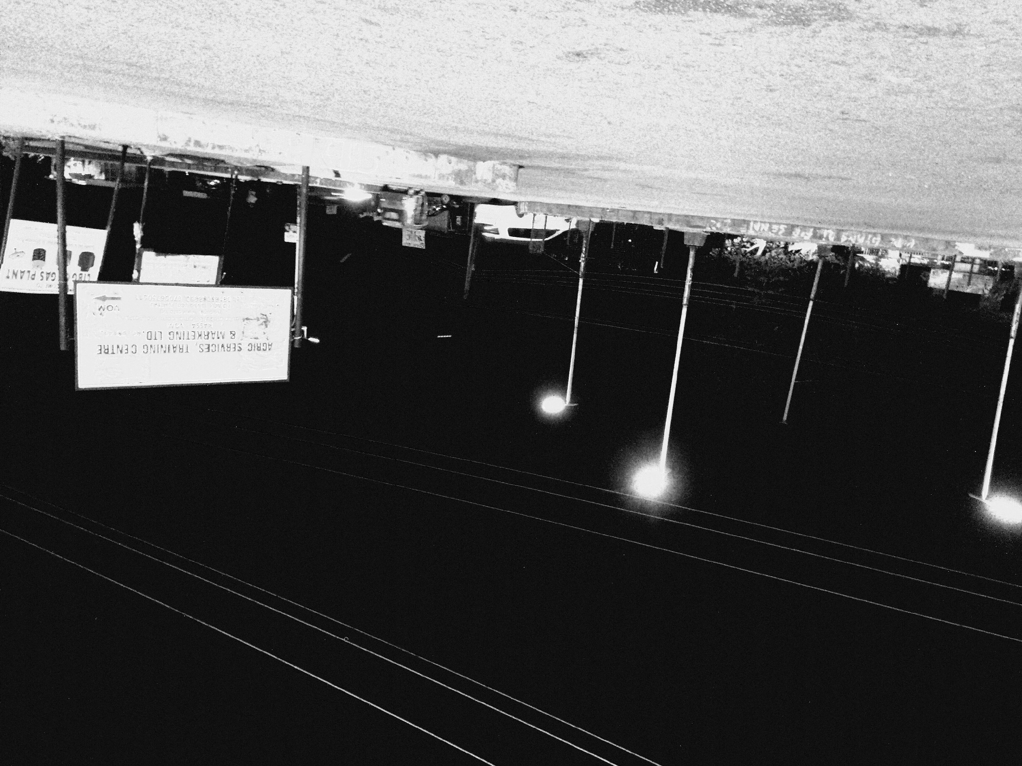 This is a black and white, night-time photograph taken on a street in Bukuru, Nigeria. In the foreground, a paved road surface is visible, with a concrete barrier along the side. Beyond the barrier, there are several tall lampposts providing illumination. In the middle ground, various vehicles are parked or driving on the road, including a white van and other smaller vehicles. There are also signs visible. One prominent sign is white with black text that reads "AGRIC SERVICES, TRAINING CENTRE & MARKETING LTD." Other signs are partially obscured, but one appears to say "GAS PLANT". In the background, the dark sky is crisscrossed by power lines. The overall scene depicts a somewhat industrial or commercial area at night, with artificial lighting illuminating the street and surrounding structures.