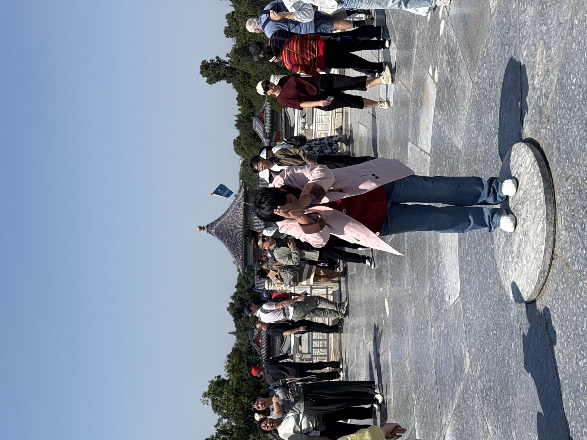 The image shows a crowd of people standing in a line at the Temple of Heaven in Dongcheng District, China. The Temple of Heaven is a complex of religious buildings dating back to the Ming and Qing dynasties. The people are mostly tourists, and some of them are taking pictures of the Hall of Prayer for Good Harvests. The Hall of Prayer for Good Harvests is a circular wooden building with a triple-eaved roof. It is one of the most recognizable buildings in the Temple of Heaven complex.
