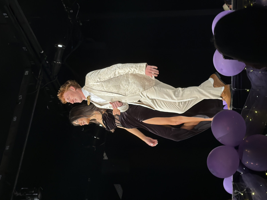Two young adults, a female and a male, are walking side-by-side across a dimly lit indoor stage, presumably in Palos Verdes Estates, United States. The female, positioned on the left, has long dark hair and wears a dark, high-slit strapless dress with a patterned midriff section, revealing her left leg up to the mid-thigh. She has a ring on her right ring finger. The male, on the right, has red hair and wears a light-colored, patterned three-piece suit (jacket, vest, trousers), a white collared shirt, and a light-colored bow tie, complemented by brown dress shoes. Both individuals are smiling and looking towards their left (viewer's left) as they walk. The stage's edge in the foreground is decorated with several purple balloons, dark netting, and small, warm-toned string lights. The background is dark, revealing overhead stage lighting fixtures and metal rigging with numbered labels (e.g., 57, 58, 59, 60, 61). A person's head and a smartphone, held upright as if recording, are partially visible in the bottom right corner, indicating an audience present.