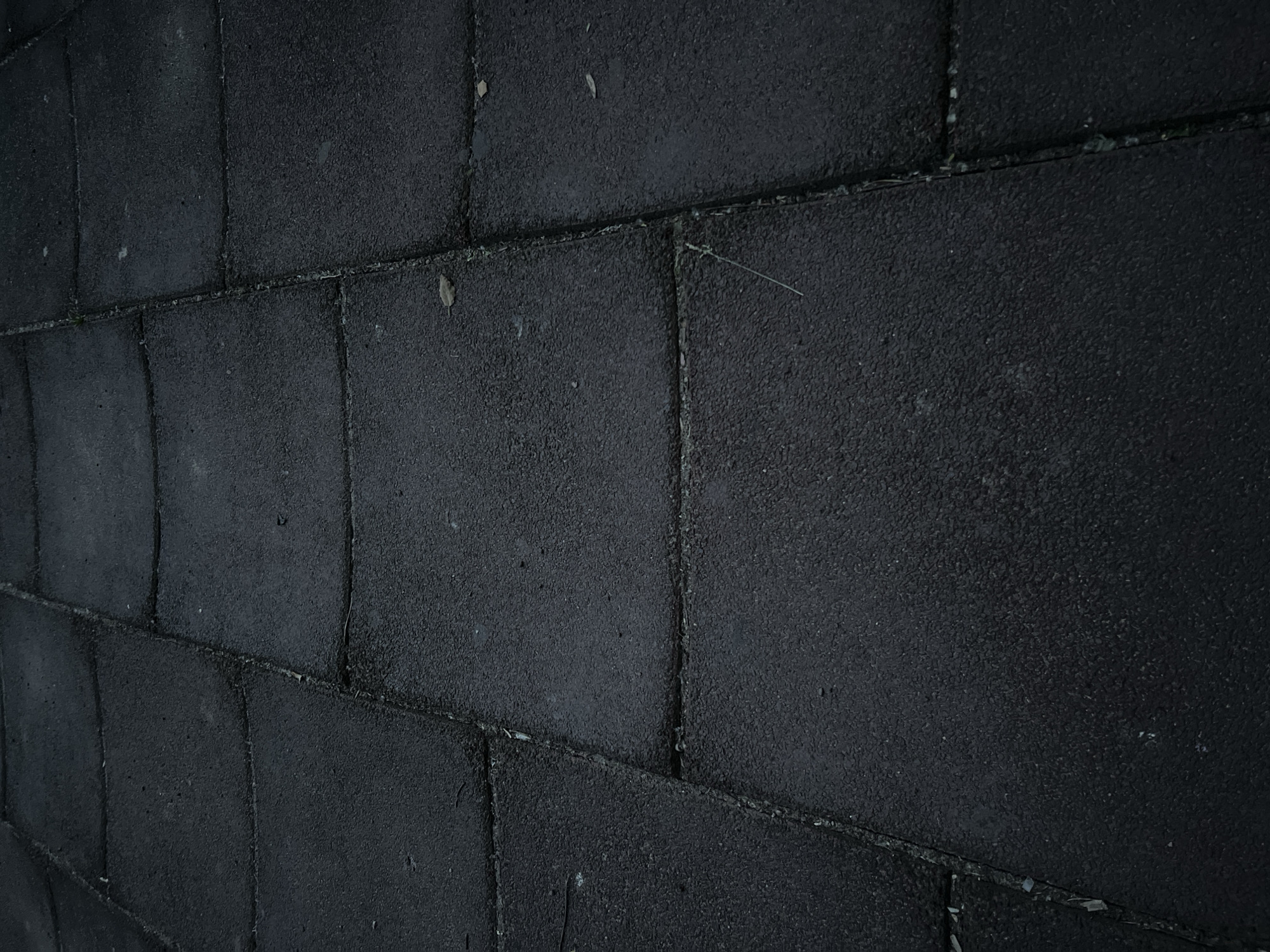 The image displays a close-up, ground-level view of a paved area constructed from dark, rectangular interlocking tiles. The tiles appear to be made of a rough, granular material, possibly rubberized asphalt or a similar composite. The surface is slightly uneven, with some tiles showing subtle depressions or warping, creating a gentle, undulating texture. The lines between the tiles are visible as thin, dark grooves, with some appearing to have accumulated a small amount of debris or sediment. A few small, light-colored particles, possibly dust or small pebbles, are scattered across the surface. A single, thin, light-colored strand, potentially a blade of grass or a thin fiber, is visible lying across one of the tile seams in the lower right quadrant of the image. The lighting is dim, suggesting either an overcast day or evening conditions, and the overall color palette is dominated by dark grays and blacks. There are no people, animals, or other significant objects present in the frame. The scene is devoid of any specific context that would indicate a particular location, and no identifiable features of the city of Sinyavino, Russia, are discernible.