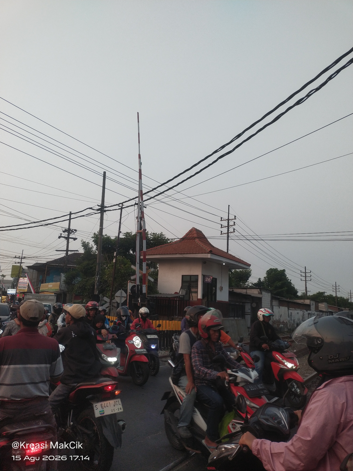 The image depicts a congested street scene, likely in Taman, Indonesia, with numerous motorcycles and riders queuing at a railway crossing. The sky is overcast and a pale gray. Dominating the mid-ground is a railway crossing signal with red and white striped poles, indicating a closed crossing. Adjacent to the crossing is a small, red-roofed building with the number "26" visible on its facade.
The foreground is filled with motorcycles and their riders. Several individuals are wearing helmets, with a variety of colors and styles visible. One rider in the foreground wears a black helmet with "DJ MARU" written on it. People are dressed in casual clothing, including t-shirts, long-sleeved shirts, and jackets. A license plate "S 6441 JCL" is visible on one of the motorcycles.
In the background, several buildings are visible, including what appears to be a shop with signage that includes the text "DOKTER HA" and "RISA DIYONGS". Power lines and utility poles are prevalent throughout the scene, stretching across the sky. The overall impression is one of a busy urban environment where traffic is temporarily halted due to the railway crossing. The timestamp "@Kreasi MakCik 15 Agu 2026 17.14" is overlaid on the bottom left corner of the image.
