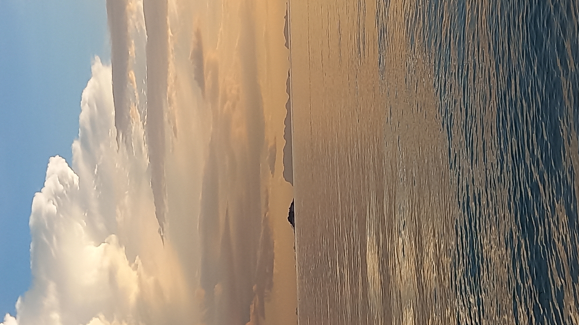 A wide shot of the ocean at sunset, with islands in the distance. The sky is filled with dramatic clouds, tinged with orange and yellow from the setting sun, contrasting with a patch of clear blue sky. The sea is calm, with gentle ripples reflecting the colors of the sky. Several islands of varying sizes are visible on the horizon, appearing as dark silhouettes against the muted sky. The scene is serene and evokes a sense of peace and natural beauty. This scene is likely from the coast of Koh Phangan, Thailand, given the reference to ตำบลเกาะพะงัน.