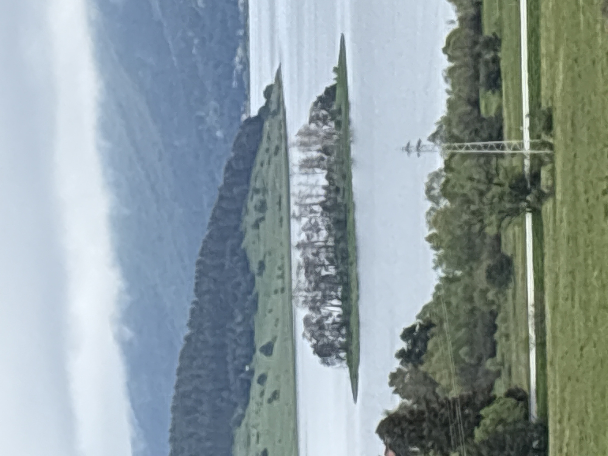 The image shows a landscape with water, likely a lake, separating two landmasses. The left landmass features a mountainside with cloud cover. The right landmass has a green grassy area and trees. An island with trees sits in the middle of the lake. A power line structure is visible on the right side of the image. The location is possibly Tsigov chark, Bulgaria. The scene is captured in overcast conditions.
