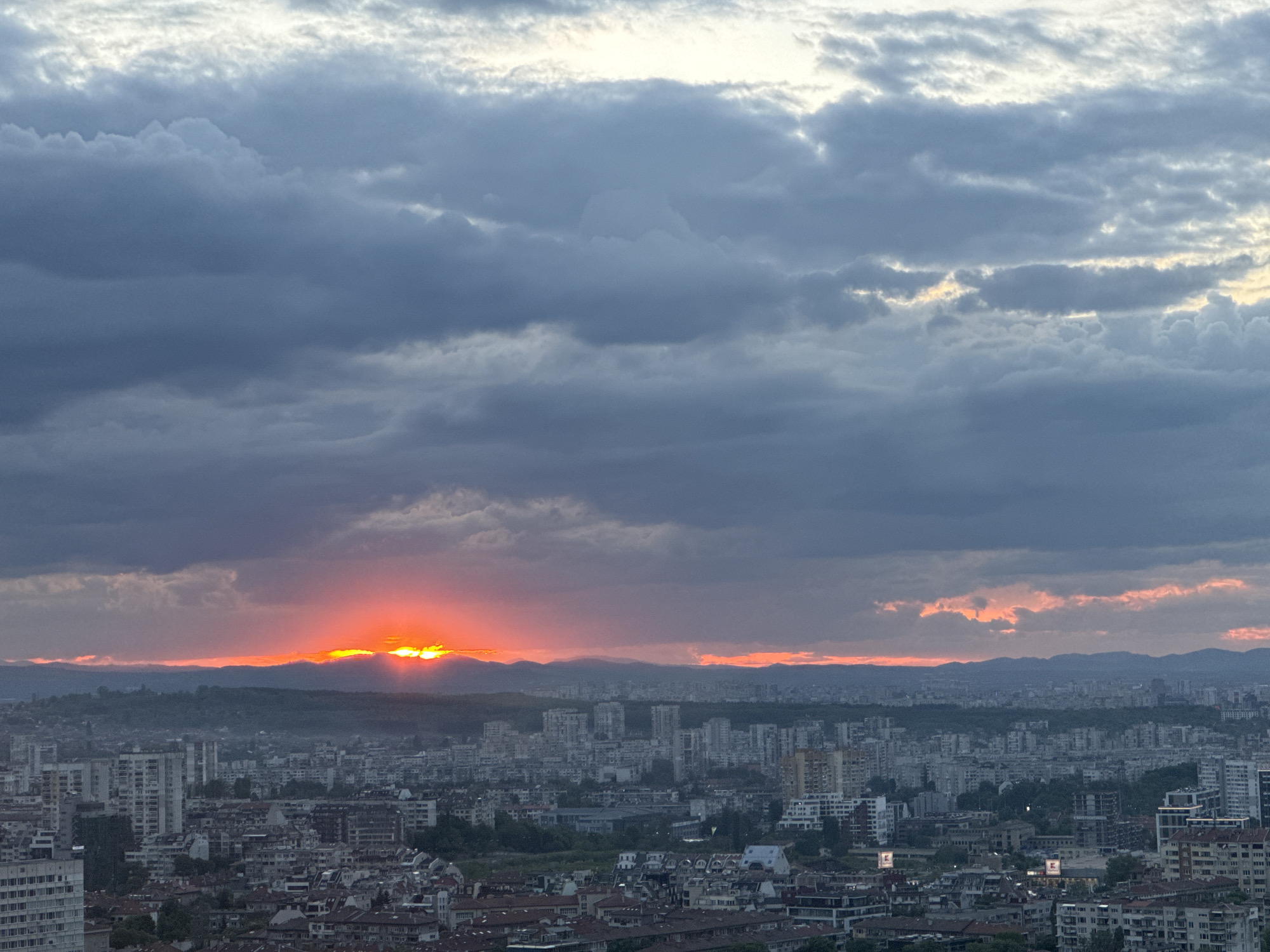 The image captures a sunset over the city of Sofia, Bulgaria. The sky is filled with layered, predominantly gray clouds with lighter patches near the top and a vibrant orange glow where the sun dips below the horizon. This glow is visible behind a distant mountain range. Below the mountains, the cityscape of Sofia is visible, featuring a dense collection of buildings, mostly residential, with a mix of low-rise and high-rise structures. A light haze or mist obscures the lower portions of the city. There are no people visible in the image. The scene is static, focused on the natural phenomenon of the sunset over an urban landscape.
