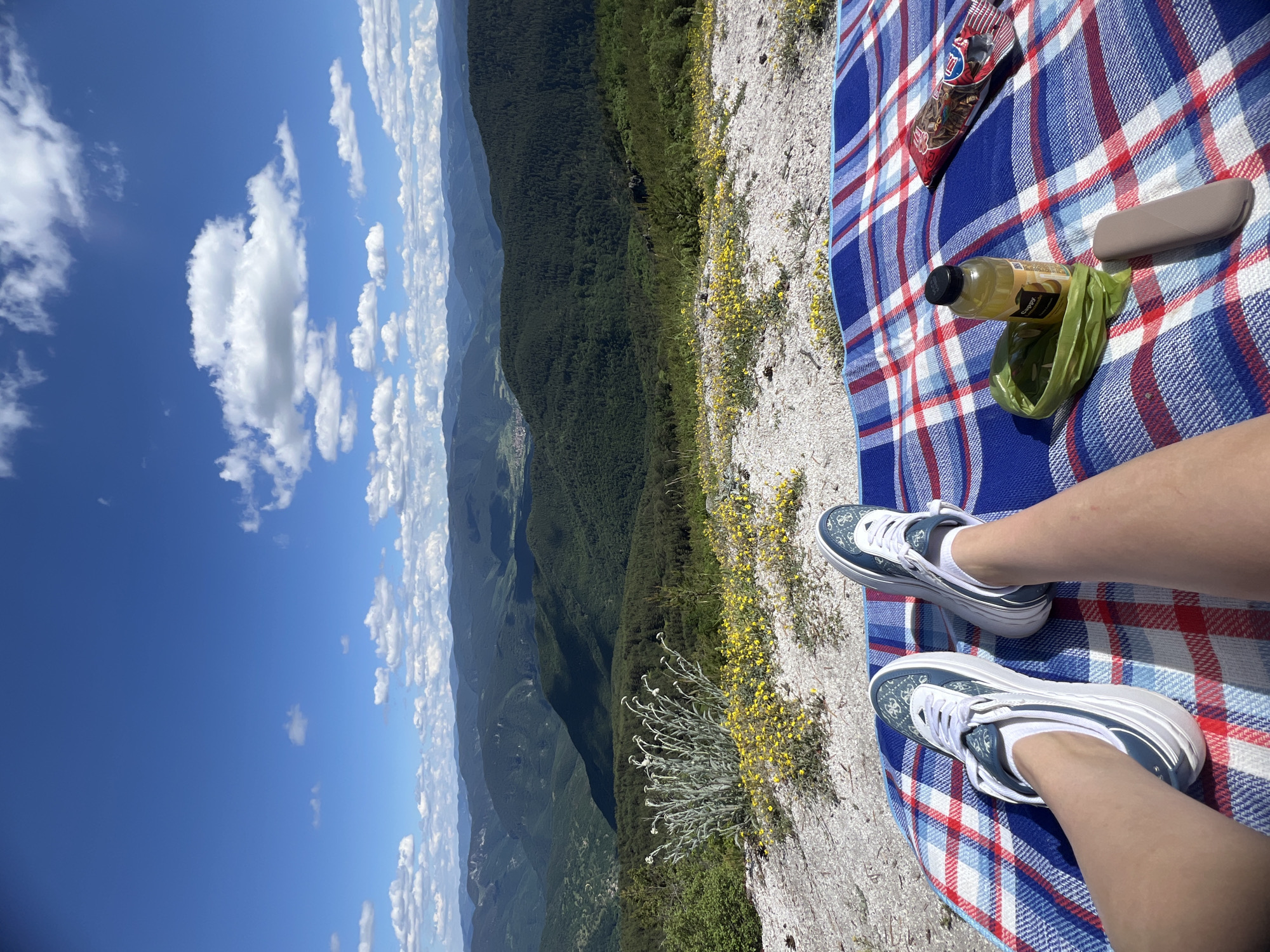 A pair of legs in white socks and blue and white sneakers rests on a blue and red plaid blanket spread on a rocky, grassy slope. Small yellow flowers dot the slope. Behind the blanket, a vast, green, mountainous landscape stretches to the horizon under a bright blue sky with scattered white clouds. A bottle of yellow liquid, a green bag, and a grey electronic device are on the blanket. A red and white package of snacks is also on the blanket. The location is identified as the city of Kosovo, Bulgaria.