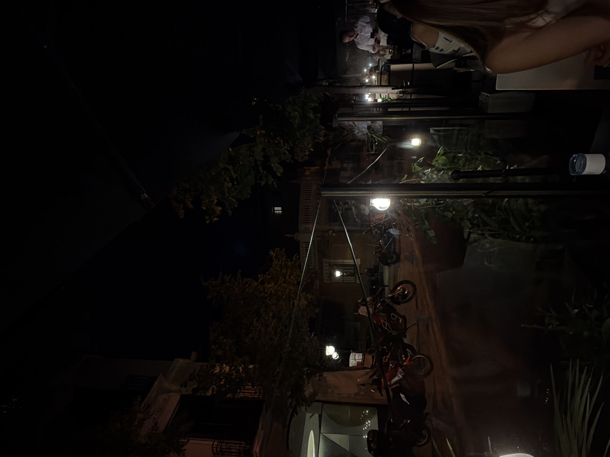 The scene is an outdoor setting at night in an urban environment in Unknown, Unknown. The area seems to be a dining establishment, as tables and chairs are visible, occupied by patrons. A woman with long blonde hair is seated at a table. A man wearing a white shirt is also present. Motorbikes are parked in the background, seemingly along a street or alley. Lighting is provided by streetlights and restaurant lighting, creating an ambiance. Vegetation, including trees and potted plants, can be seen.