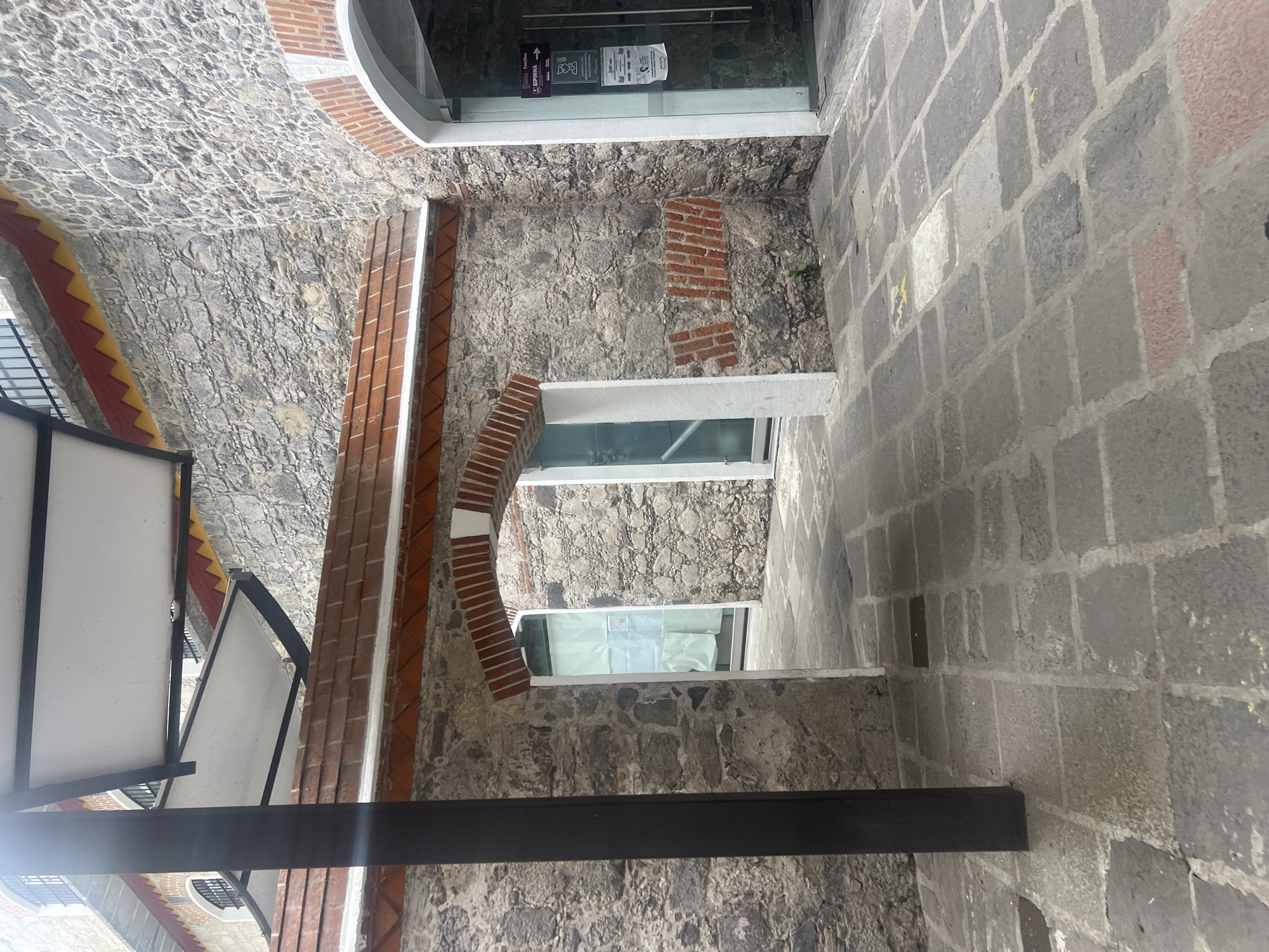 This eye-level shot captures a narrow passageway in Puebla, Mexico, constructed from roughly hewn gray stones. The walls are also constructed from stone. Brick accents are visible around arched openings and along sections of the walls. A dark, possibly metal, beam crosses the foreground horizontally. Windows, visible on the wall, are covered. Signage is visible at the top of the frame. The passage opens to what appears to be an exterior space.