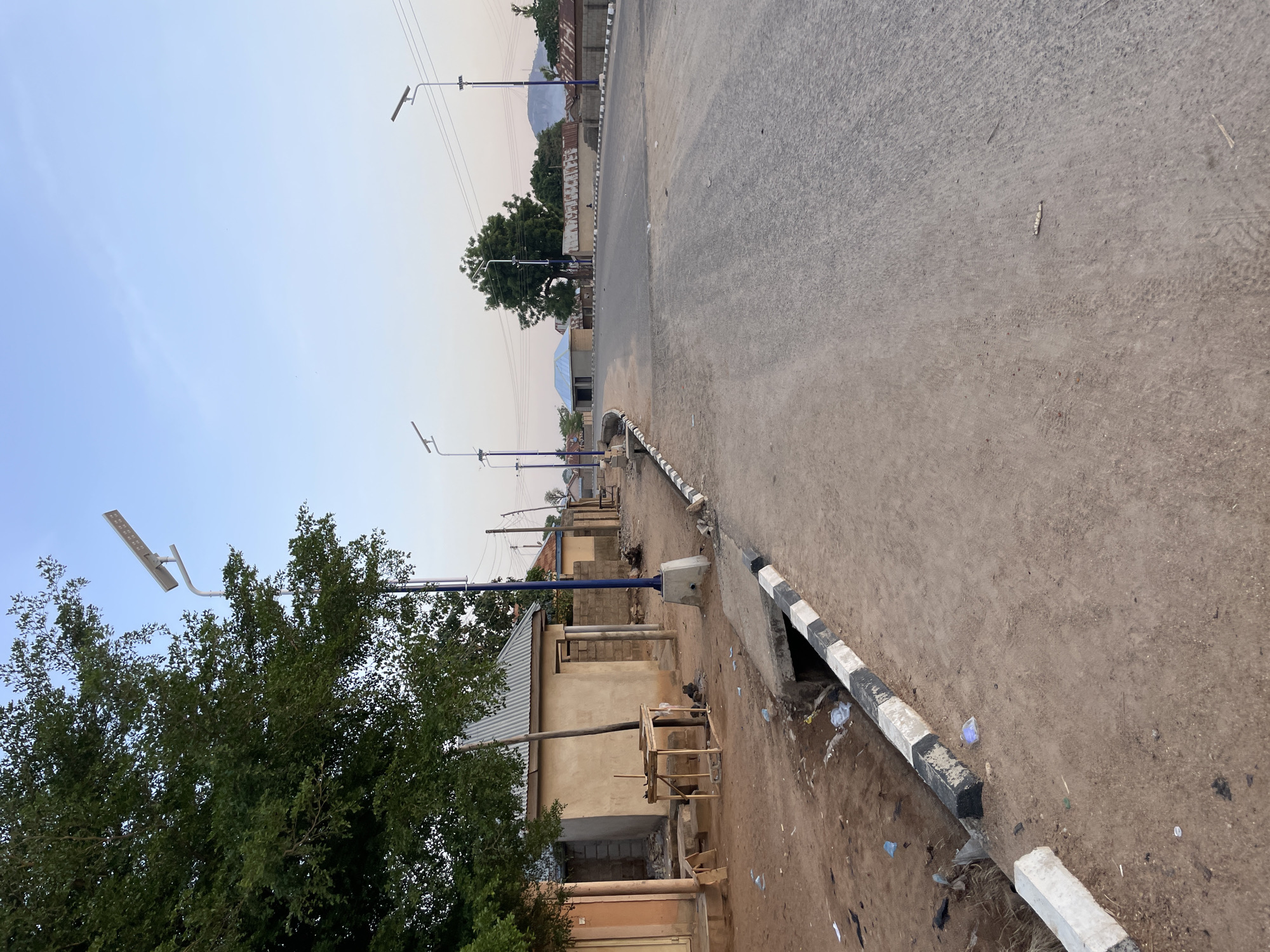 The image depicts a street scene in Bauchi, Nigeria. The road is paved and stretches into the distance, occupying the right side of the frame. A striped black and white curb runs along the edge of the road on the right. To the left of the curb is a dirt area littered with trash. Adjacent to the dirt area are buildings of varying construction, some with corrugated metal roofs. A large tree is visible on the left side of the frame. Streetlights with solar panels are spaced along the road, on the left side. Overhead power lines are also visible. In the distance, buildings and a hill are visible under a light sky. The scene is devoid of people.