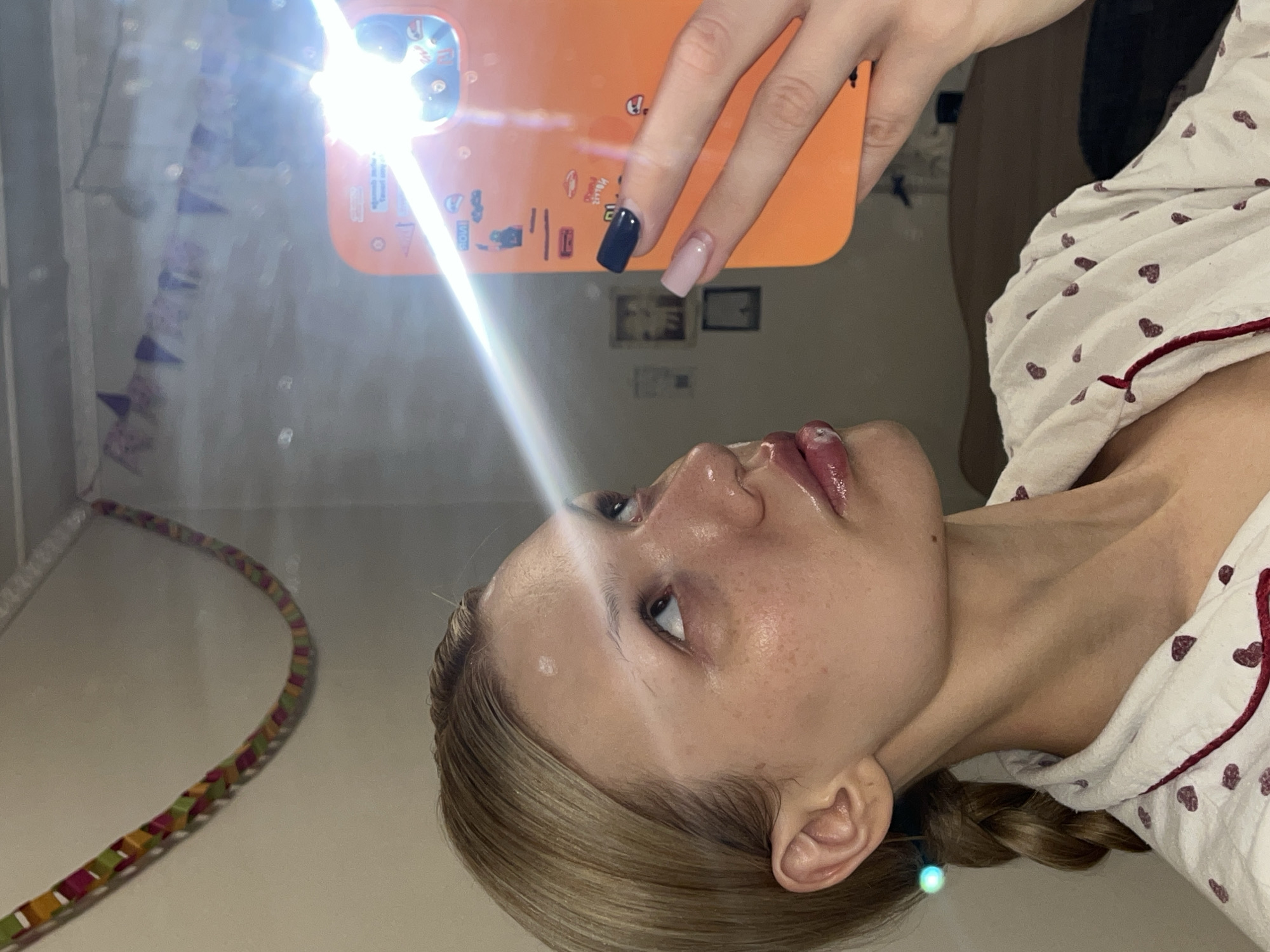 A woman is holding a phone, presumably taking a selfie. She has blonde hair, with a visible braid. She is wearing pajamas with a heart print. The phone is orange. In the background, there are what appear to be paper decorations hanging on the wall. The scene is indoors. The location in the city of Chaikovsky, Russia is not discernible from the image.