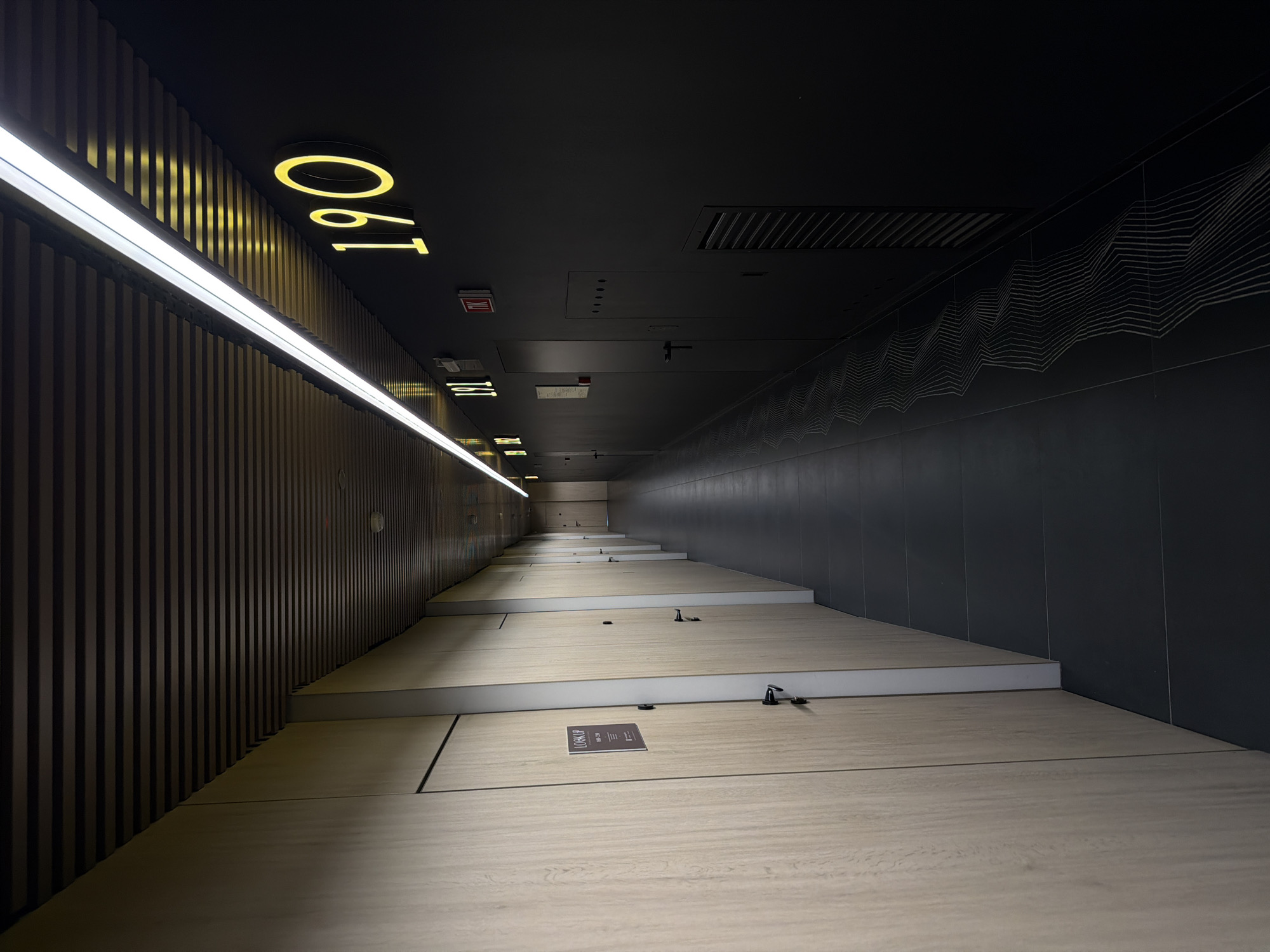 This is an interior shot of a hallway in Moscow, Russia. The hallway is lit by a long, linear light fixture on the left-hand side. The numbers "190" are displayed on the wall near the light fixture. The floor is made of large, light-colored rectangular tiles. There are black metal fixtures attached to the floor. The right-hand wall is dark gray with a linear, wavy pattern near the ceiling. The ceiling is dark and features several recessed panels and vents. The hallway stretches into the distance with progressively smaller versions of the floor tiles and wall elements.