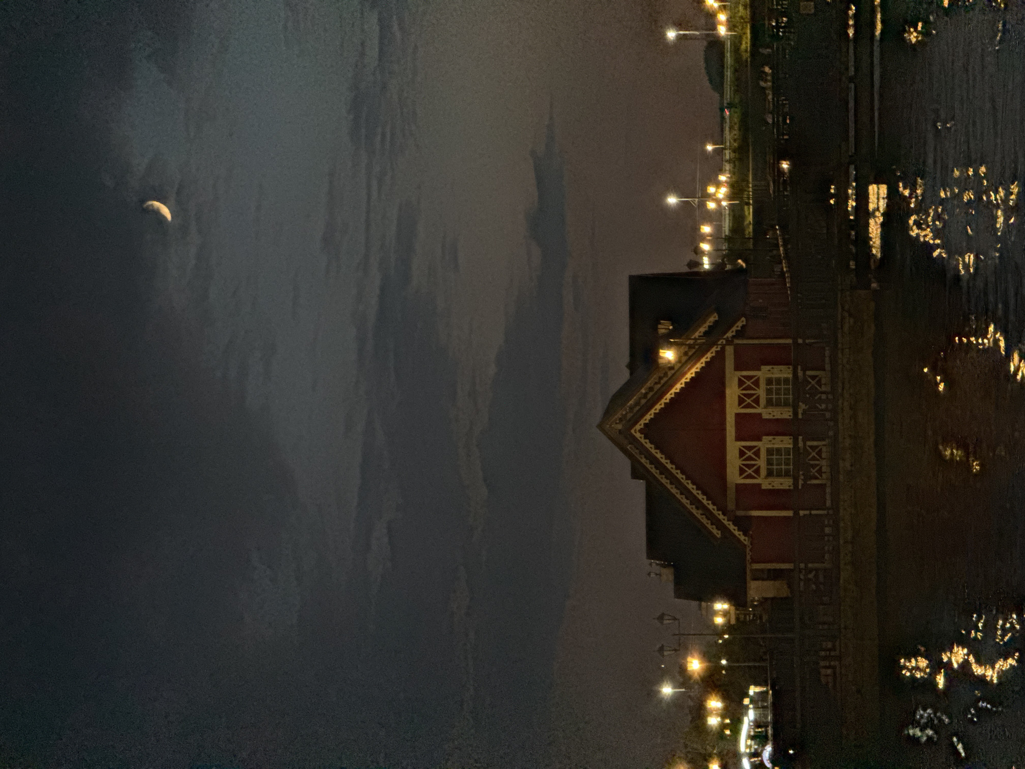 The image depicts a nighttime scene at a waterfront in Kazan, Russia. A red wooden building with white trim and decorative windows is centrally located, bordering a body of water. The water reflects the lights from nearby streetlamps, creating a shimmering effect. In the sky, a crescent moon is visible amidst a cloudy, dark expanse. The environment appears to be a public space with a walkway or deck in front of the building and rows of streetlights lining the background.