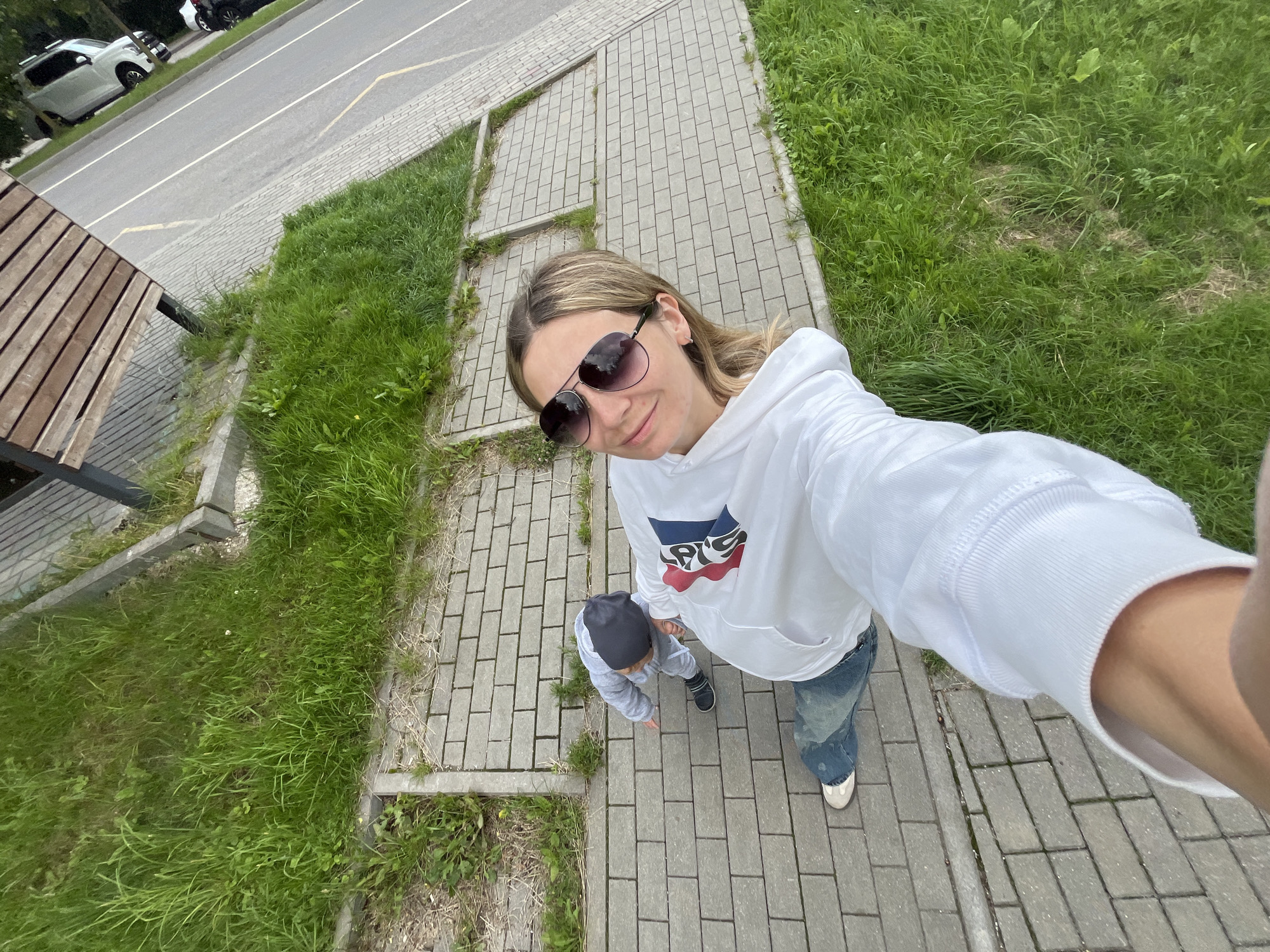 A woman wearing a white hoodie and blue jeans, with large sunglasses, takes a selfie on a sidewalk. Beside her, a young child in a gray tracksuit and black beanie walks on the sidewalk. The sidewalk is made of grey rectangular pavers and is bordered by a strip of green grass. Beyond the grass, there is a paved area with a wooden bench and a street with a white car parked. The scene appears to be in an outdoor urban environment, possibly a park or a residential area. The background suggests a street with a white van and trees, indicating a location within the city of Pionersky, Russia.