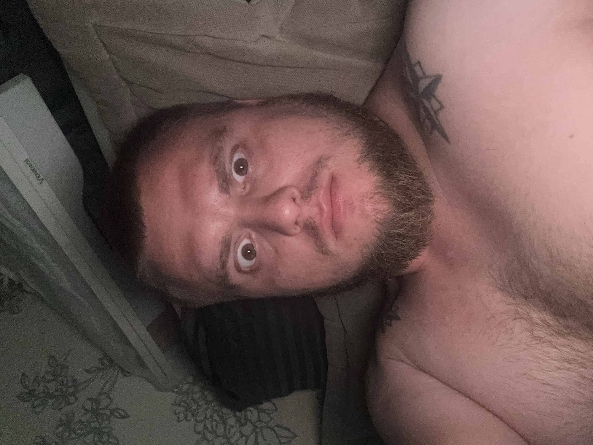 The image shows a close-up, selfie-style photograph of a shirtless man with a beard, lying down and looking directly at the camera with wide eyes. He is positioned on what appears to be a couch or bed, with a textured fabric background. To the left of the man, there is a dark pillow and a wall with a floral wallpaper pattern. Above the man's head, a light-colored object with the word "VENERDI" printed on it is visible, suggesting it might be electronic equipment or a piece of furniture. The man has tattoos on his shoulder and chest. The lighting is dim, casting a reddish hue across his skin. There are no discernible landmarks or elements that would definitively place this image within Pyatigorsk, Russia.