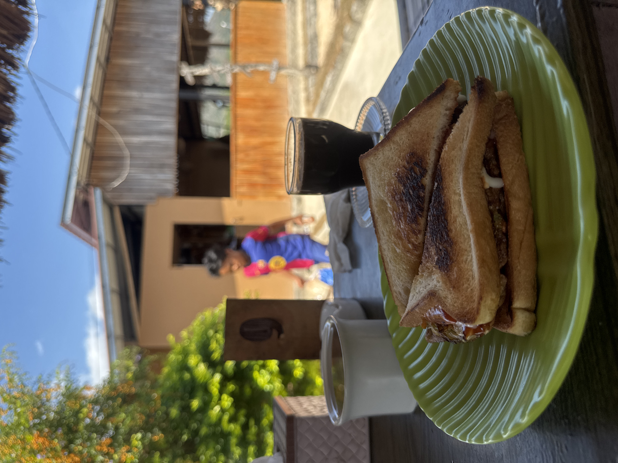 In Kertasari, Indonesia, a close-up view of a breakfast is captured at a roadside eatery. In the foreground, a light green, wavy-rimmed plate holds two halves of a toasted sandwich, filled with what appears to be meat and sauce. To the left of the sandwich is a white mug, possibly containing tea, and to the right, a clear glass filled with a dark liquid, likely coffee, rests on a clear saucer. Behind the table, a wooden sign with the number "9" is visible, and a child wearing a blue and red shirt walks in the background. The eatery has a rustic, open-air design with wooden paneling and a thatched roof visible at the top of the frame. Lush green foliage and a blue sky with scattered clouds complete the outdoor setting.