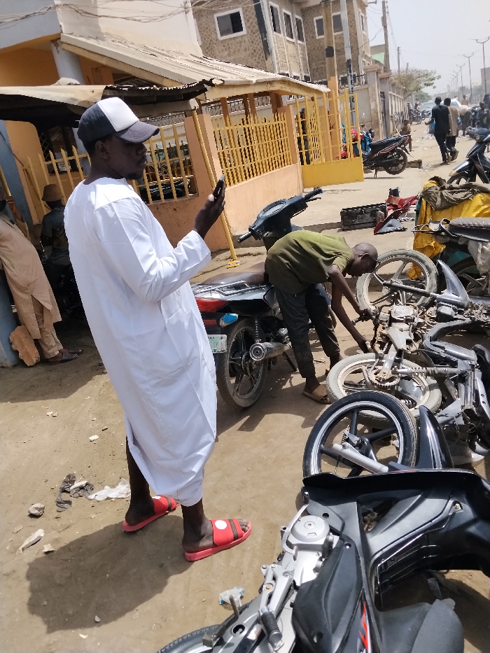 The scene depicts an outdoor urban environment in Fagge D2, Nigeria, under bright daylight. In the immediate foreground and slightly to the left, a dark-complexioned adult male stands, wearing a white long-sleeved tunic, a grey and white baseball cap, and red slide sandals. He holds a black smartphone vertically with both hands, looking down at the screen.
To his right, in the mid-ground, another dark-complexioned adult male, wearing an olive-green shirt and dark pants, is bent over, actively engaged in repairing or disassembling a motorcycle engine or frame. This individual wears light-colored open-toed footwear. Numerous motorcycle components, including frames, wheels, and an engine block, are scattered across the dusty, unpaved ground in the foreground and mid-ground, indicating a motorcycle repair or salvage area. A black motorcycle with a silver exhaust is parked upright directly behind the man in the white tunic. A partially disassembled black motorcycle occupies the bottom right corner of the frame.
In the background, a single-story building with a corrugated metal roof and a prominent yellow metal gate or fence structure is visible to the left. Further back, a two-story building with a textured, stone-like facade and multiple windows stands. Distant figures are observable walking along what appears to be a street, and several more motorcycles are parked or positioned further down the road. Debris is present on the unpaved ground.