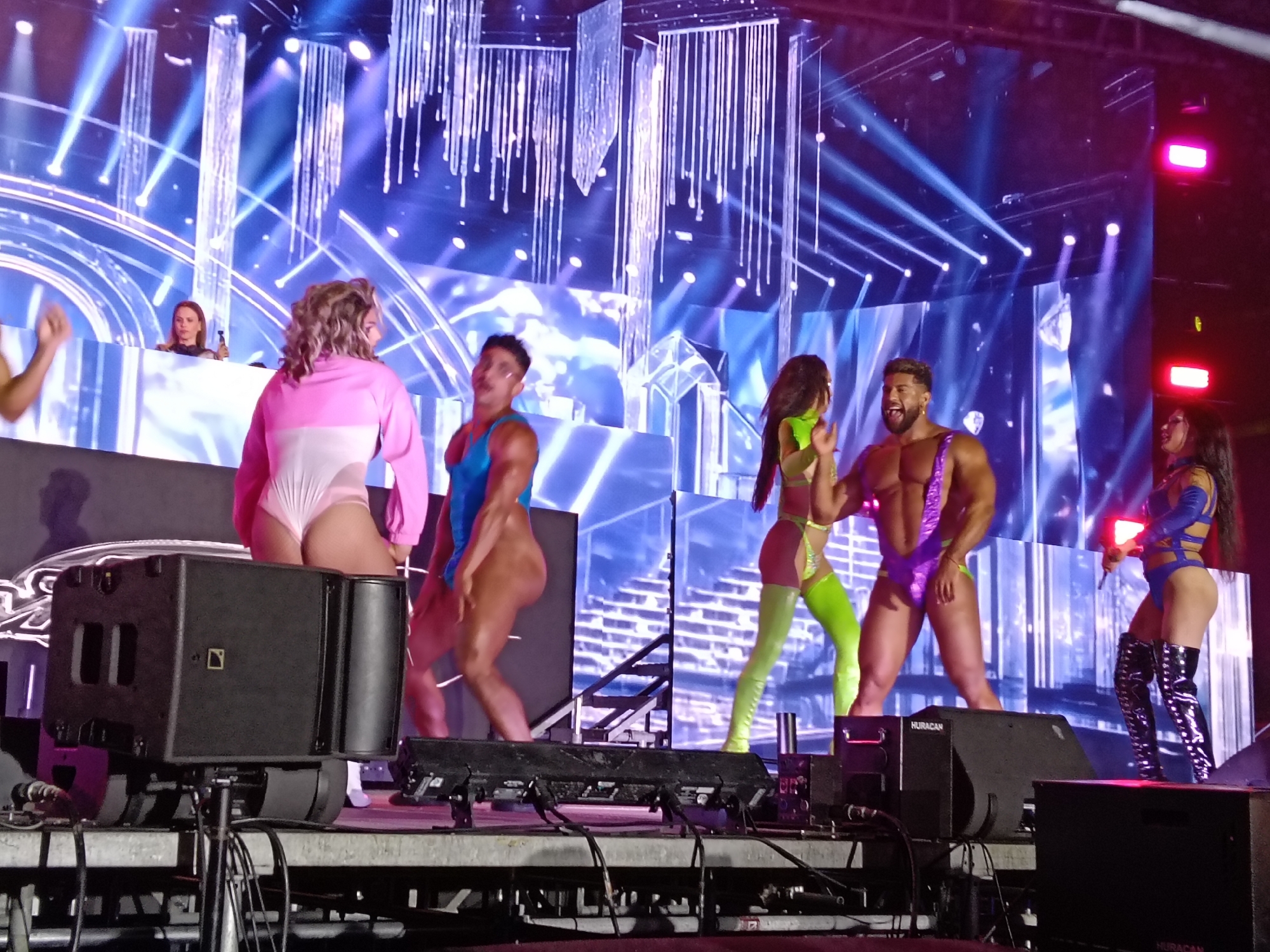 The image captures a stage performance, likely a concert or show, in San Bartolomé de Tirajana, Spain. Several performers are visible, dressed in revealing, colorful outfits. The backdrop is a complex arrangement of lights and digital displays. Stage equipment, including speakers marked "HURACAN," is prominent in the foreground. In the background, a DJ booth with a woman visible suggests a musical performance. The setting appears to be an outdoor venue or a large indoor space set up for a performance.