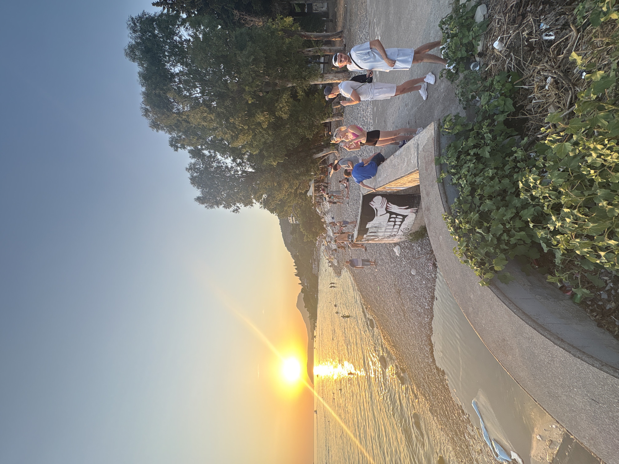 This image depicts a beach scene at sunset in Bar, Montenegro. The sun is low on the horizon, casting a warm glow over the calm sea and the pebble beach. Several people are visible along the shoreline, some wading in the water and others sitting or standing on the beach. In the foreground, a concrete wall with graffiti runs along the edge of the promenade, where a group of four individuals is standing and observing the sunset. Trees line the promenade behind the group, and a hilly landscape is visible in the distance. The overall atmosphere is serene, with the golden light of sunset creating a tranquil mood.