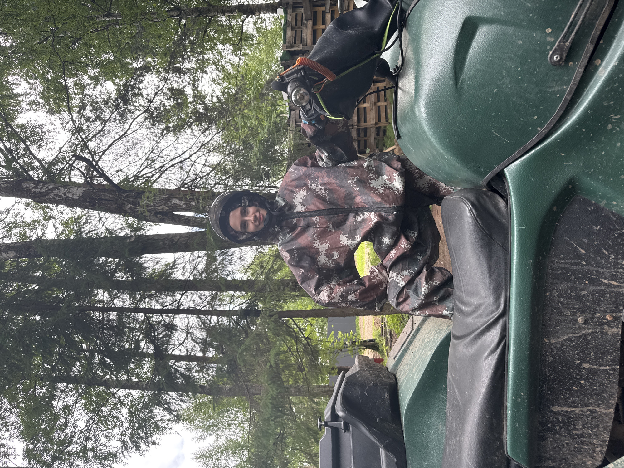 A young adult individual, appearing female, is positioned next to a mud-splattered green all-terrain vehicle (ATV) in a wooded outdoor environment. The individual wears a full-body camouflage suit with a pixelated pattern of brown, white, and dark grey tones, and a black helmet is worn over their head. A faint smile is present on their face as they look directly at the viewer. Their left hand grips the left handlebar of the ATV, which features a black handguard and an orange coiled cord accessory. The ATV has a black seat and a black storage box mounted on the front. A small, bright yellow object is visible near the individual's right hip. The background consists of dense coniferous and deciduous trees under an overcast sky. A structure constructed from wooden pallets is visible on the right side of the background, and a dark fence is partially visible further back. The ground is dirt, indicating an off-road setting. The precise location within the city of Perm, Russia, cannot be determined from the visual information provided in the image.