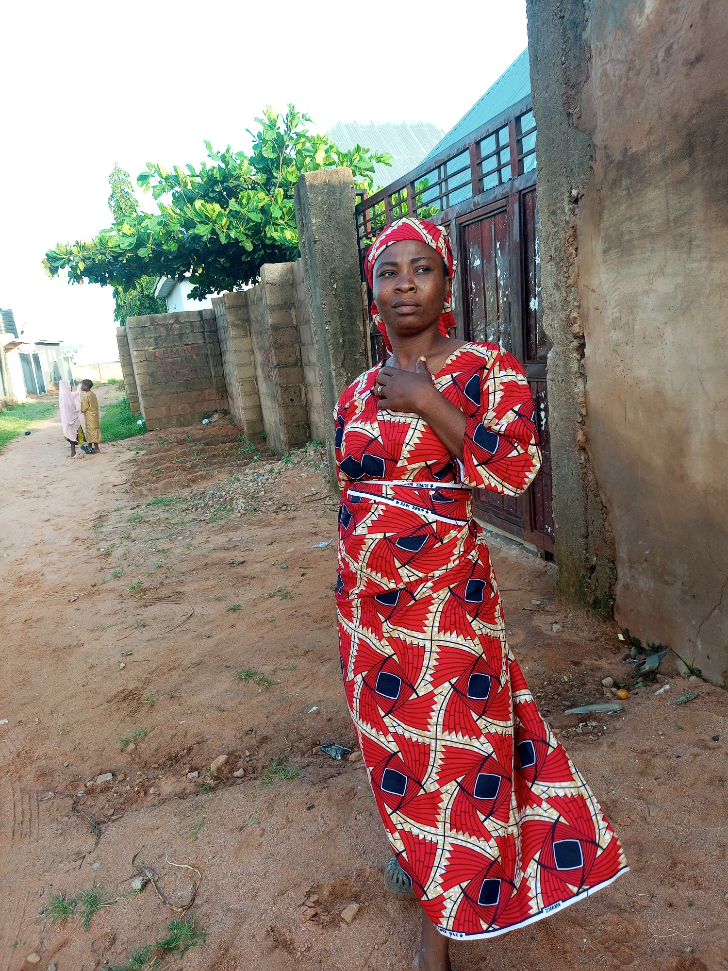 In Tempure, Nigeria, a woman stands near a rough-textured wall and a weathered metal gate. She wears a vibrant red, white, and black patterned dress and a matching headscarf. Her left hand rests on her chest, and she looks to her left with a thoughtful expression. In the background, a dirt path leads towards a cluster of small buildings. Two young children, one in a pale pink outfit and the other in yellow, walk away from the camera down the path, partially obscured by a brick wall and a lush green tree. The ground is sandy and uneven, with scattered debris.