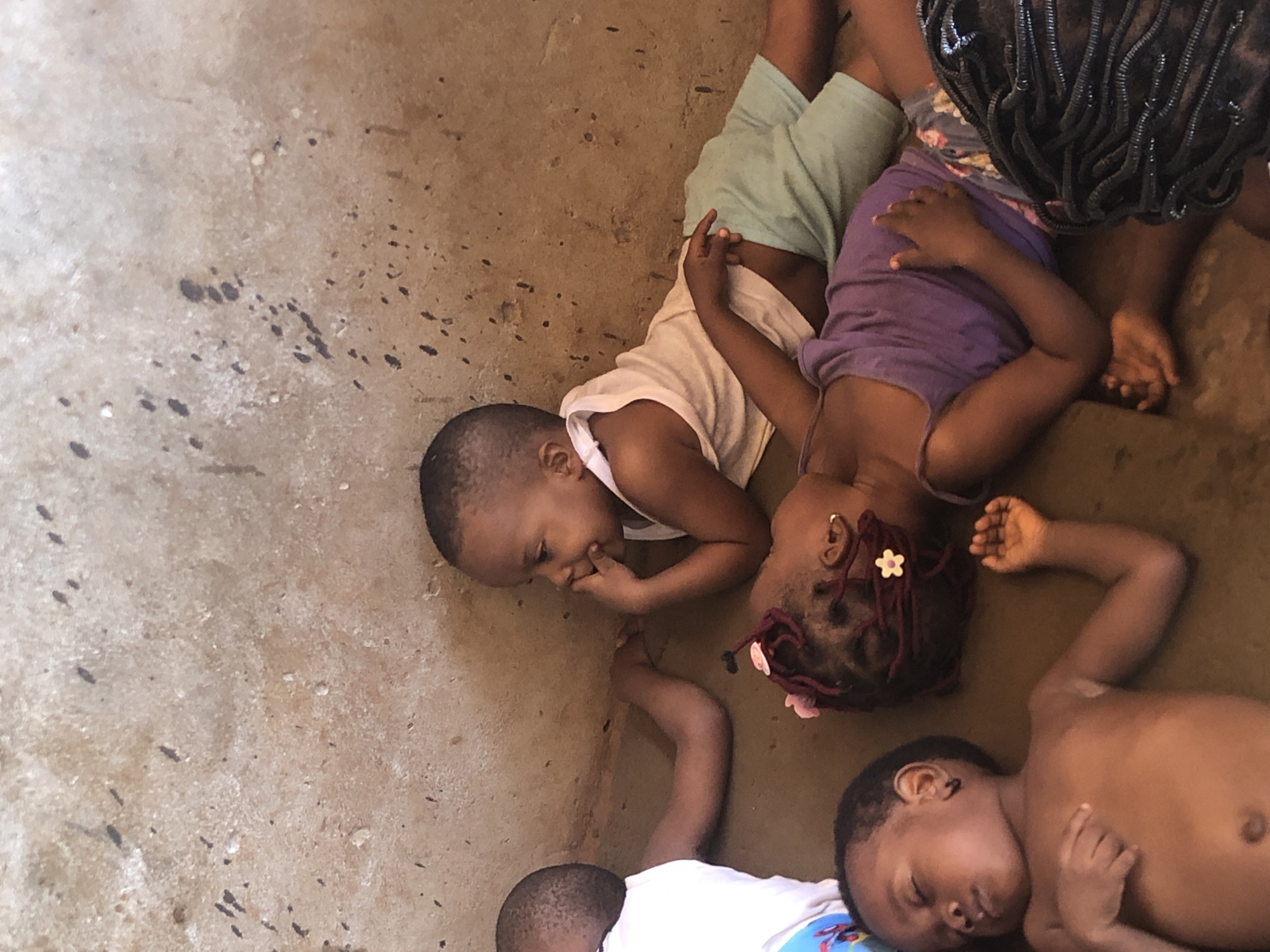 The image shows three young children lying on a brown floor in what appears to be a room in Ukwu-Abwa, Nigeria. The floor has dark splatter marks. Two of the children are awake and lying beside each other. The first child is lying on their stomach, wearing a white tank top and light green shorts, looking towards the center of the image with their hand near their mouth. The second child is wearing a purple top and has braided hair adorned with two white and pink hair accessories. They appear to be looking at the first child. A third child is lying near the other two; they are sleeping on their back, shirtless, with closed eyes. The top of the image contains a section of a person's head with dark braided hair. Their right hand is resting on the sleeping child's arm.