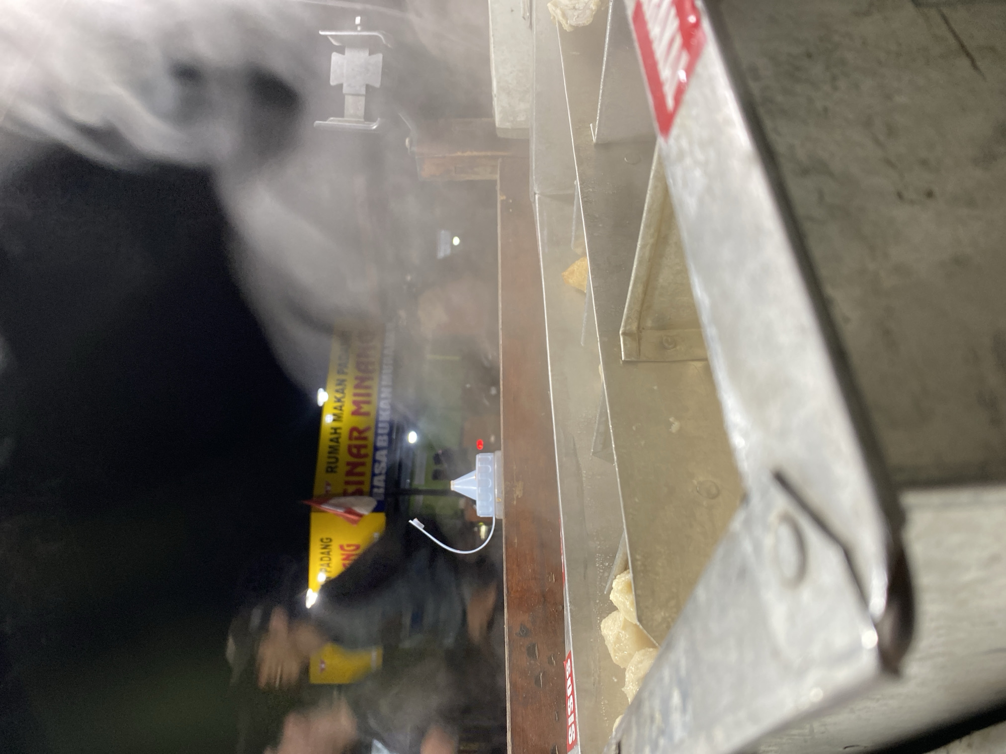 In Kasihan, Indonesia, a night scene unfolds at a food stall. The foreground is dominated by a metal food preparation area, with compartments containing food items that appear to be light-colored meatballs or dumplings. A red and white sticker with illegible text is visible on the edge of the preparation area. Behind this, a wooden counter is present, topped with a white plastic sauce bottle with a blue cap and a white flexible tube. In the background, a yellow sign with red and black text reads "PADANG" and "SINAR MINANG" suggesting it is a Padang restaurant. Blurred figures of people are visible behind the counter, with one person wearing a cap. Steam rises from the food, obscuring some of the background elements and creating a hazy effect. The lighting is dim, consistent with an outdoor night setting.