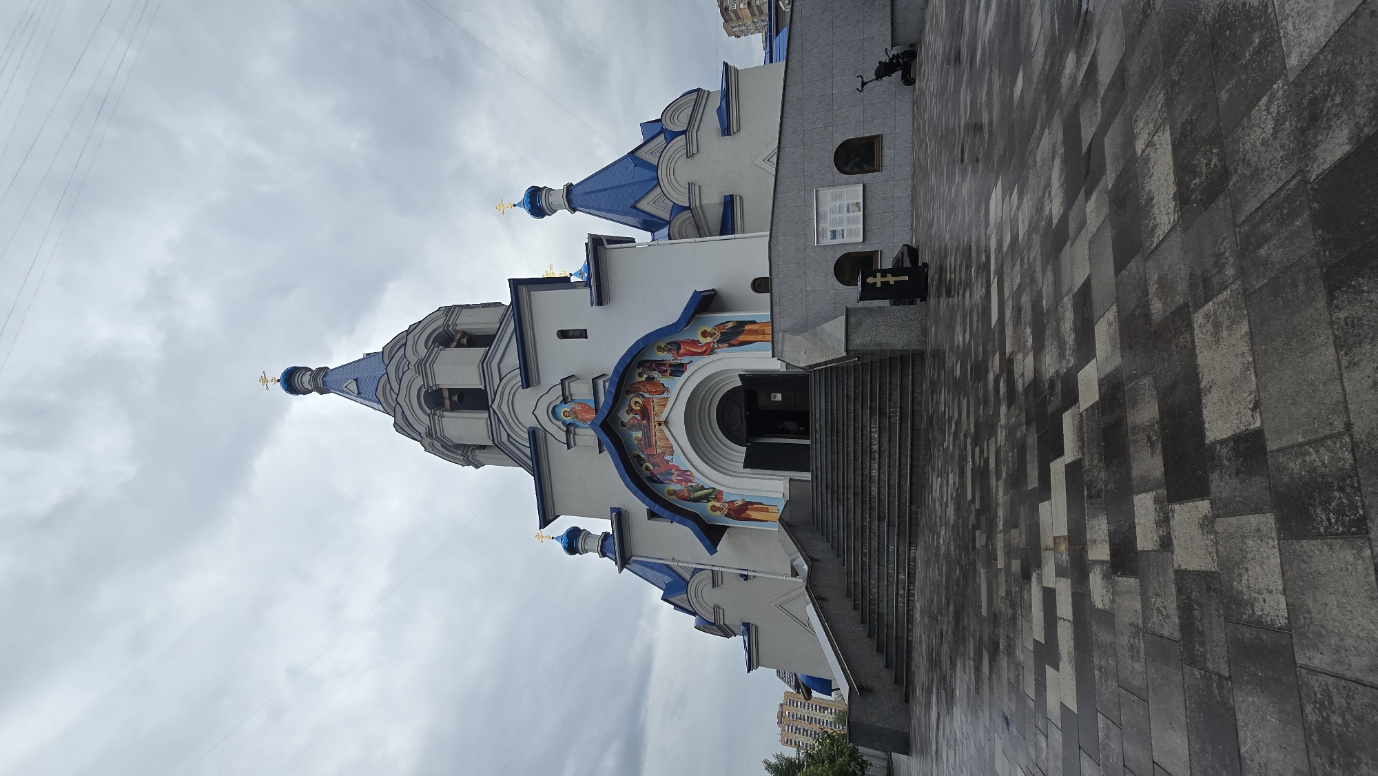 This full, eye-level, slightly low-angle exterior shot depicts the Church of the Intercession of the Blessed Virgin Mary, located at vul. Mykilsko-Slobidska, 4A, in Kyiv, Ukraine, under an overcast sky. The prominent structure is a multi-domed Orthodox Christian church with light grey walls and distinctive blue-tiled roofs and onion domes, each topped with a golden Orthodox cross.
A wide, multi-tiered concrete staircase with grey handrails leads directly to the church's main arched entrance. Above the entrance arch, a colorful religious mural depicts various figures and scenes. The building features additional smaller blue domes and a bell tower section with arched openings visible on the upper levels.
The foreground consists of a patterned paved surface, alternating between light and dark grey rectangular blocks, visibly wet from recent precipitation, reflecting the grey sky. To the right of the staircase, a lower section of the church's facade is clad in grey textured tiles, featuring two small arched windows and a white notice board containing documents. A black, rectangular stand with a prominent gold Orthodox cross is positioned on the pavement near the base of the stairs. Further to the right, a dark stroller or similar wheeled object is partially visible. To the left, a small section of a green tree and a light-colored fence are visible.
The sky above is uniformly grey and cloudy, with thin overhead utility wires traversing the upper frame. In the distant background, on both the left and right sides, glimpses of multi-story residential buildings are visible, indicating an urban setting. A few indistinct, blurry figures appear near the church entrance, suggesting minimal human presence.