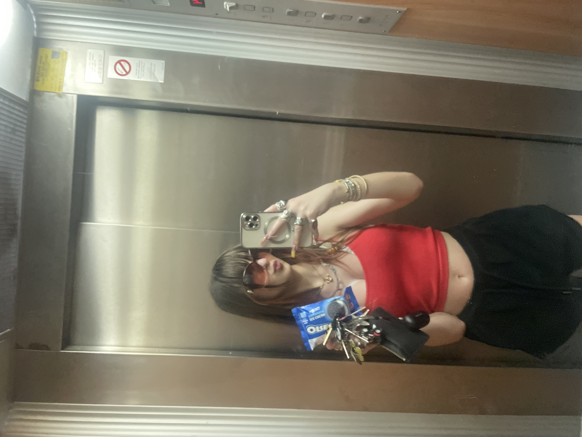 A person is taking a selfie in what appears to be an elevator. The elevator's interior is stainless steel. There is an indicator panel above, with buttons and floor indicators. Warning signage is visible on the upper section of the elevator door. The person taking the photo is wearing a red top, a black skirt, and sunglasses. They are holding an iPhone in their left hand and a pack of Oreo ice cream sandwiches with keys in their right hand. The person is wearing multiple rings and bracelets. There is no indication of the location of Petah Tikva, Israel, in the image.
