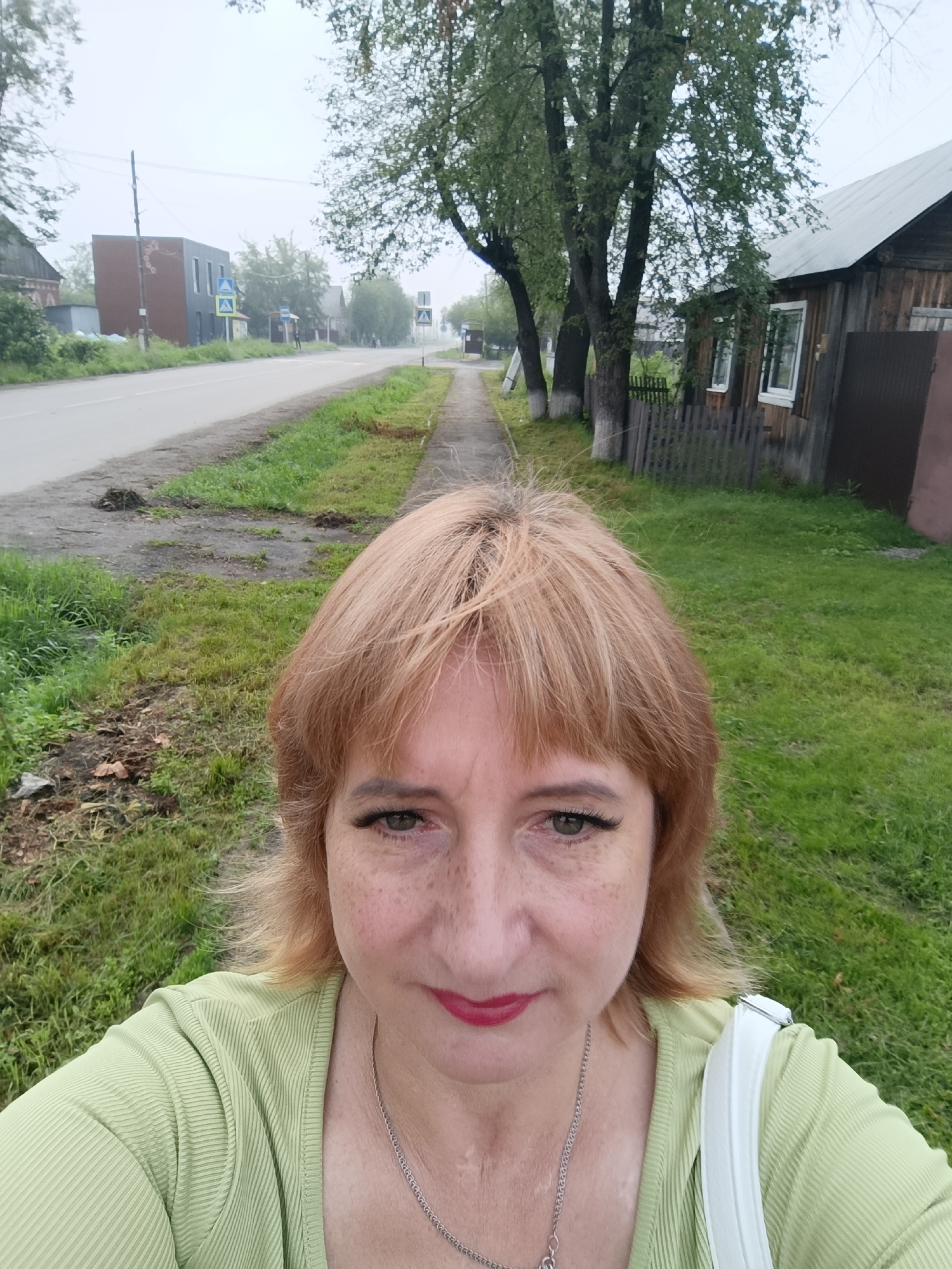 A middle-aged Caucasian woman with blonde hair and freckles is taking a selfie outdoors. She is wearing a light green ribbed shirt and a silver necklace. She has makeup on, including mascara and red lipstick. She is standing on a sidewalk next to a grassy area. In the background, there is a paved road with traffic signs and some buildings, including a wooden house with a fence. The environment appears to be a somewhat overcast day in a rural or semi-rural area, likely in Tugulym, Russia, given the architectural style of the buildings and the overall atmosphere.