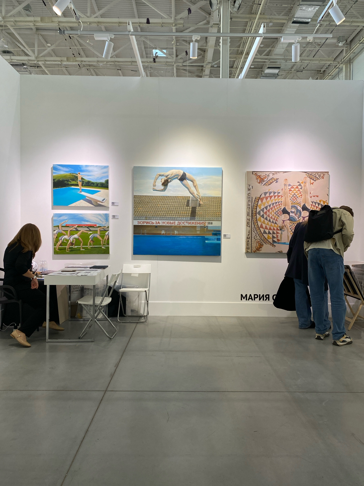 The image depicts an art exhibition space, possibly located within the city of Khimki, Russia, judging by the Cyrillic text visible. The scene features a white wall displaying several pieces of artwork, predominantly paintings with a recurring theme of athletic figures, particularly divers and gymnasts. One painting shows a person on a diving board by a pool, another displays gymnasts in a field, and a third depicts a diver in mid-air over a pool, with Cyrillic text that appears to read "БОРИС ЗА НОВЫЕ ДОСТИЖЕНИЯ" (Boris for new achievements). A fourth artwork is a mosaic-style piece featuring figures, possibly divers, with Cyrillic text encircling it.
In the foreground, a woman with dark hair, wearing a black top and pants, sits at a white table, engaged with some papers. Two white folding chairs are placed near the table. To the right, two individuals, one in a light-colored jacket and jeans, and another in a dark top and jeans, are observing the artworks. The floor is a polished concrete surface. Overhead, a track lighting system illuminates the exhibition. The overall environment suggests a public gallery or exhibition hall, with a minimalist aesthetic.