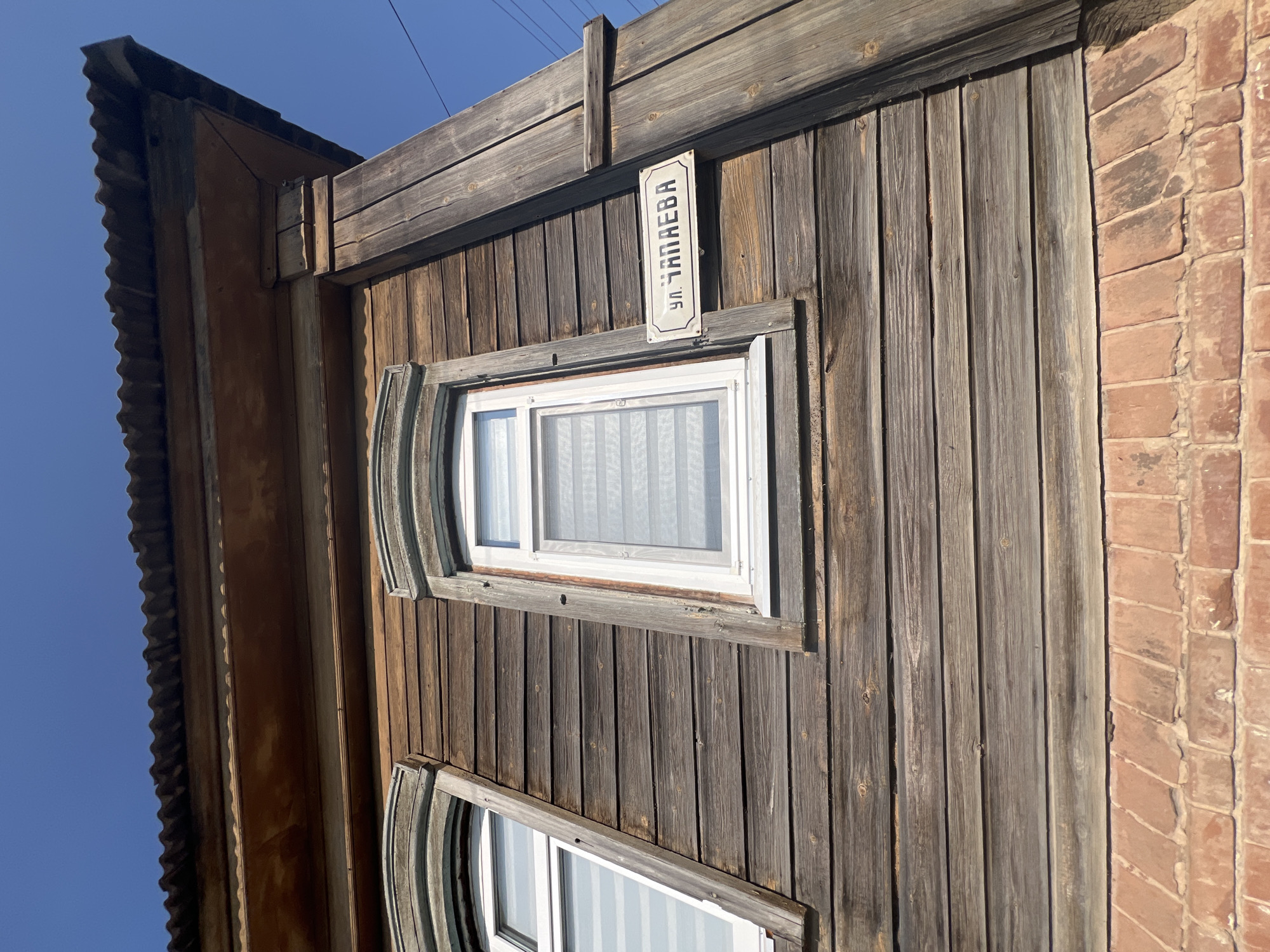 The image shows the facade of an old wooden house in Balakovo, Russia. The house is constructed of horizontal wooden planks, and two windows with white frames are visible. Above the windows, there are decorative carved wooden elements. A street sign with Cyrillic text, "ул. ЧАПАЕВА", is attached to the wooden wall. The roof of the house is covered with corrugated metal. The lower part of the house is made of red brick. The sky is clear and blue. The image appears to be taken from a low angle, looking up at the building.