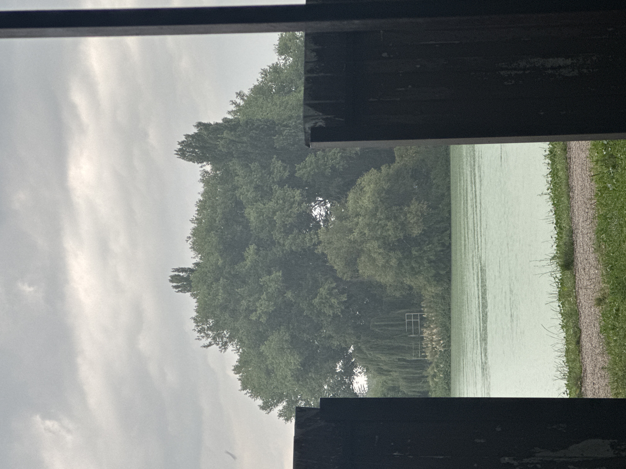 The image depicts a view through a narrow opening between two dark, vertical structures, likely wooden posts or fence panels. In the foreground, there is a gravel and grass path. Beyond the path is a body of water with ripples on its surface. The water appears to be a light green color. Across the water, a dense group of trees with lush green foliage dominates the background. Within the trees, a structure with a light-colored railing is partially visible. The sky above is overcast with heavy gray clouds. The scene is likely outdoors and appears to be a natural landscape, possibly near a lake or river. There are no people or animals visible in the image, and no specific Yessentuki landmarks are identifiable.