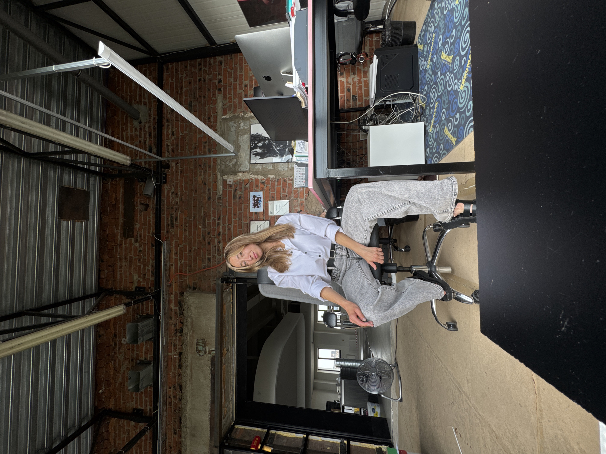 A woman sits in an office chair in a loft-style workspace. She has blonde hair and is wearing a white button-down shirt, a black belt, and light gray wide-leg jeans. The room has exposed brick walls, a metal ceiling with exposed beams and pipes, and industrial-style lighting. To her right is a desk with computers and office supplies. In the background, there is a large open doorway leading to another part of the building, where a white, undulating ceiling structure is visible. A floor fan is placed to the left of the woman, and a large black desk or table is in the foreground, partially obscuring the bottom of the image. The floor appears to be unfinished concrete or a similar material, with a patterned rug underneath the desk on the right. The overall impression is of a modern, unfinished office or creative space.