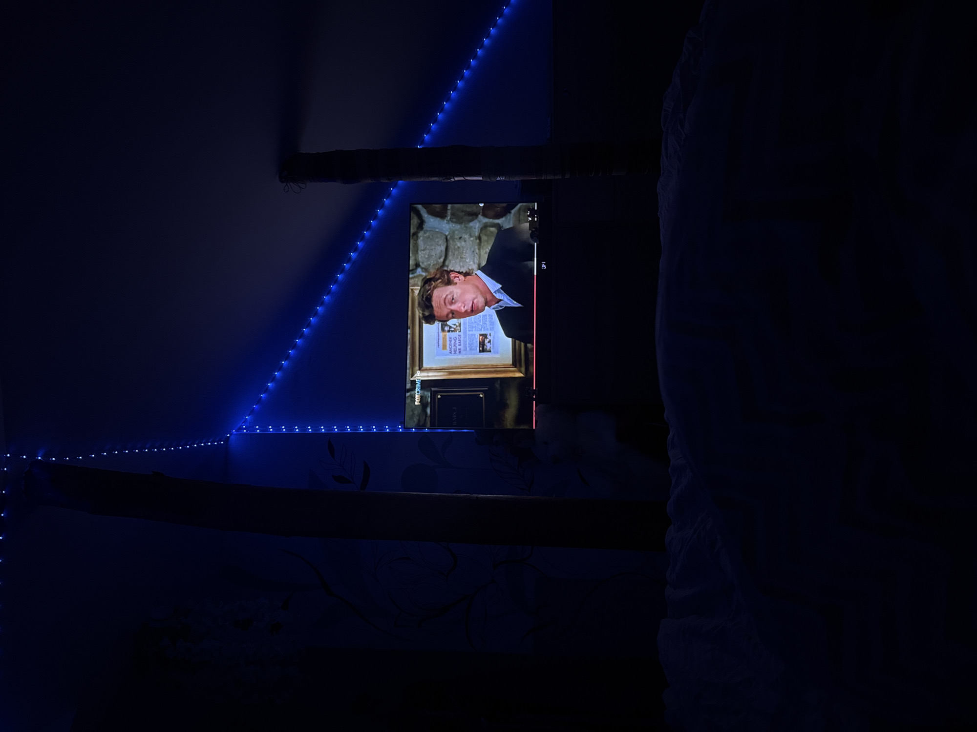A dimly lit room with blue LED strip lights along the ceiling and wall. A television displays a man in a suit, possibly from a show or movie. A wooden beam is partially visible on the ceiling. The scene appears to be indoors, possibly a bedroom, with a bed visible on the right. No specific details indicate the location is Elhovo, Bulgaria.