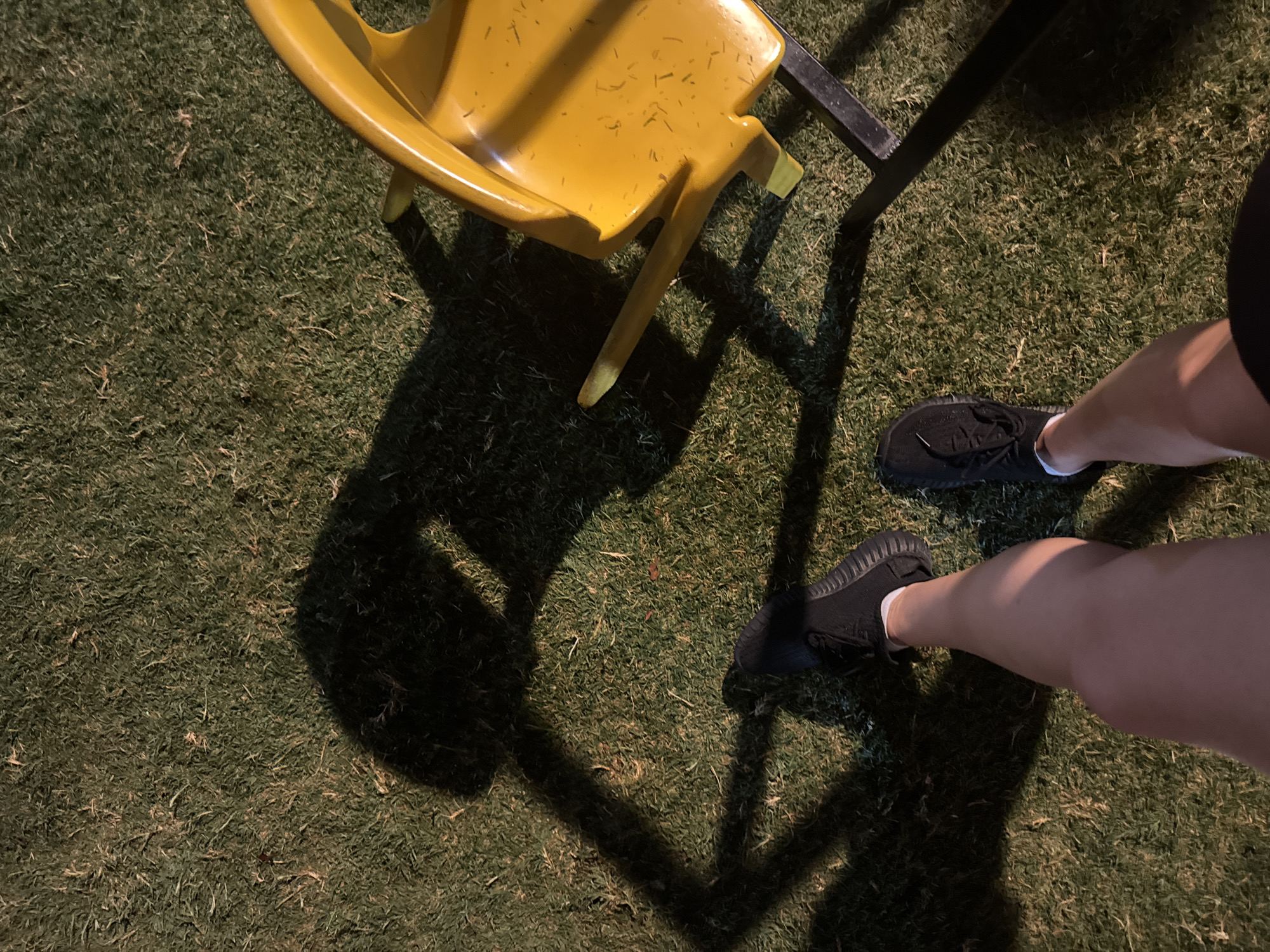 A first-person perspective shot shows a person standing on a grassy area, possibly a park or garden, with a shadow cast from overhead lighting, creating a dark, elongated silhouette on the ground. In the mid-right of the frame, a bright yellow plastic chair with a metal base is partially visible. The person is wearing black athletic shoes with white socks. The overall impression is of an outdoor scene at dusk or night, with artificial light illuminating the grass and casting strong shadows. There is no identifiable information in the image that would suggest a specific location in Aksu, Turkey.