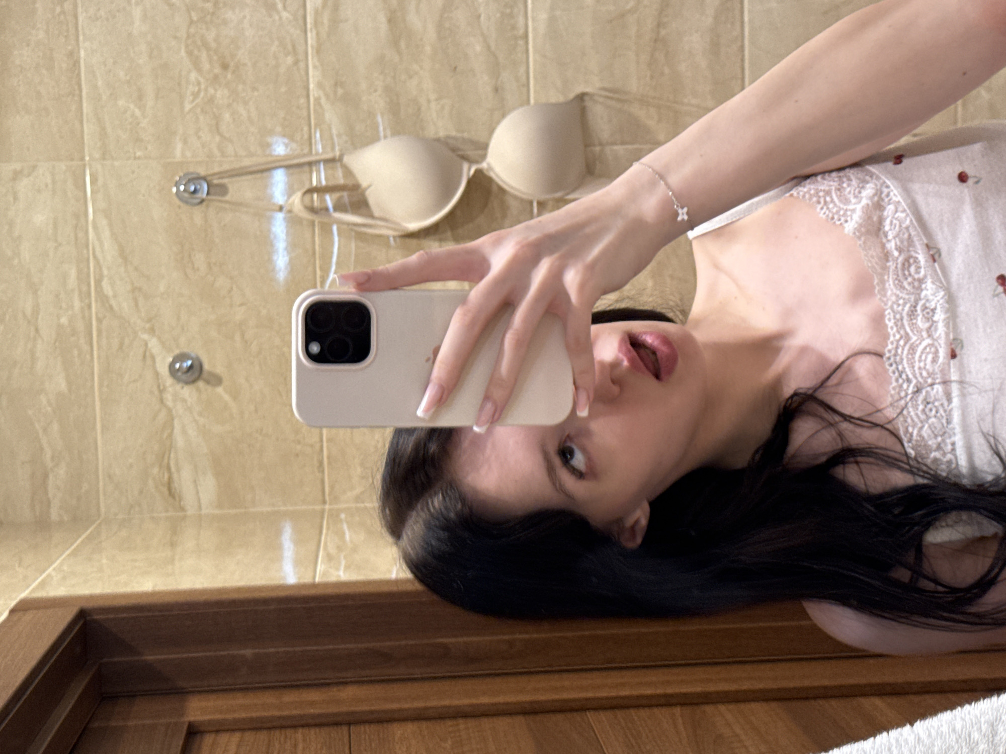In Khimki, Russia, a young woman with long, dark hair takes a selfie in a bathroom. She is wearing a white camisole with a lace trim and a cherry pattern. Her right hand, holding a cream-colored smartphone, is raised to capture the reflection. A delicate silver bracelet with a star charm adorns her left wrist. Behind her, a beige bra hangs on a hook against a tiled wall. To the left of the frame, a portion of a wooden door is visible. The bathroom walls are tiled with beige marble.